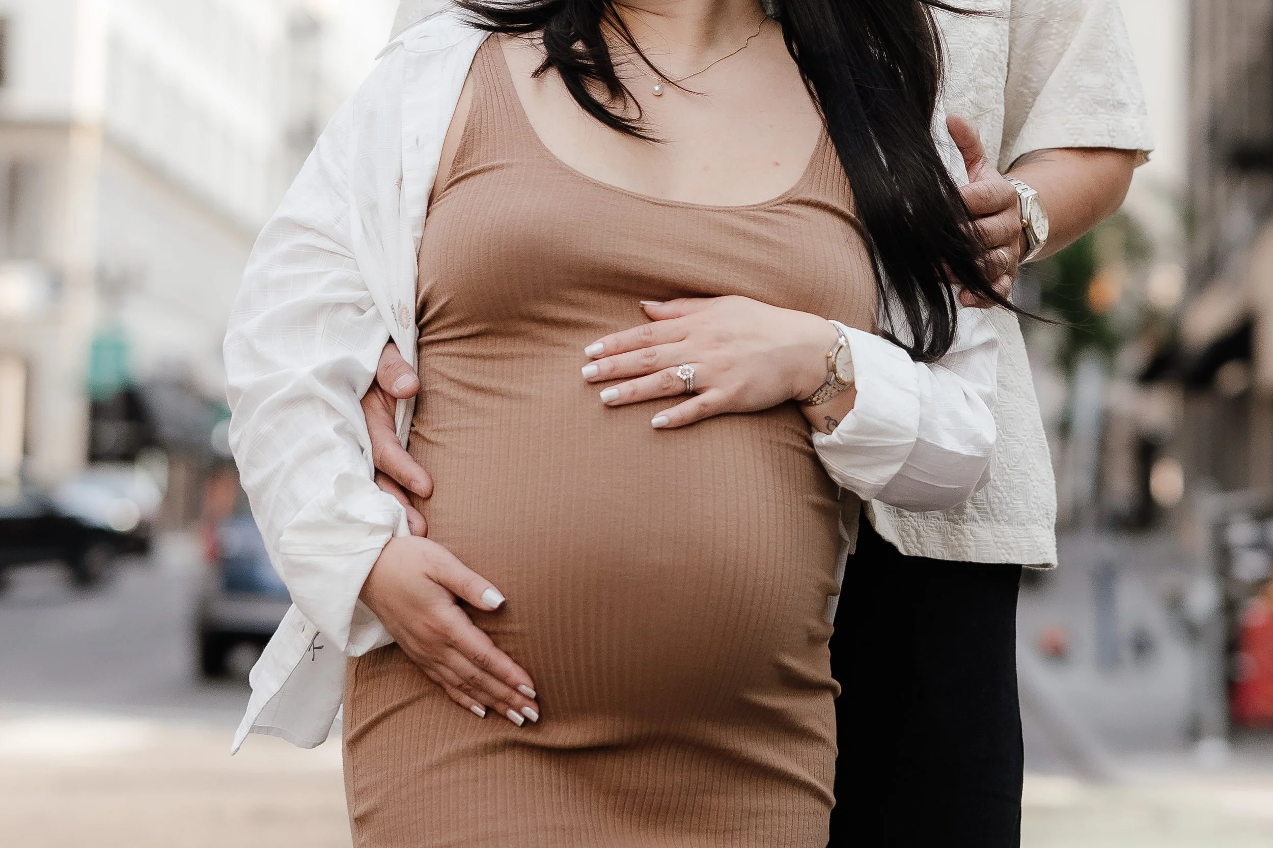 Close-up of a pregnant woman in a brown ribbed dress with a white shirt unbuttoned over it, being embraced from behind by an adult, likely a partner, on a city street.