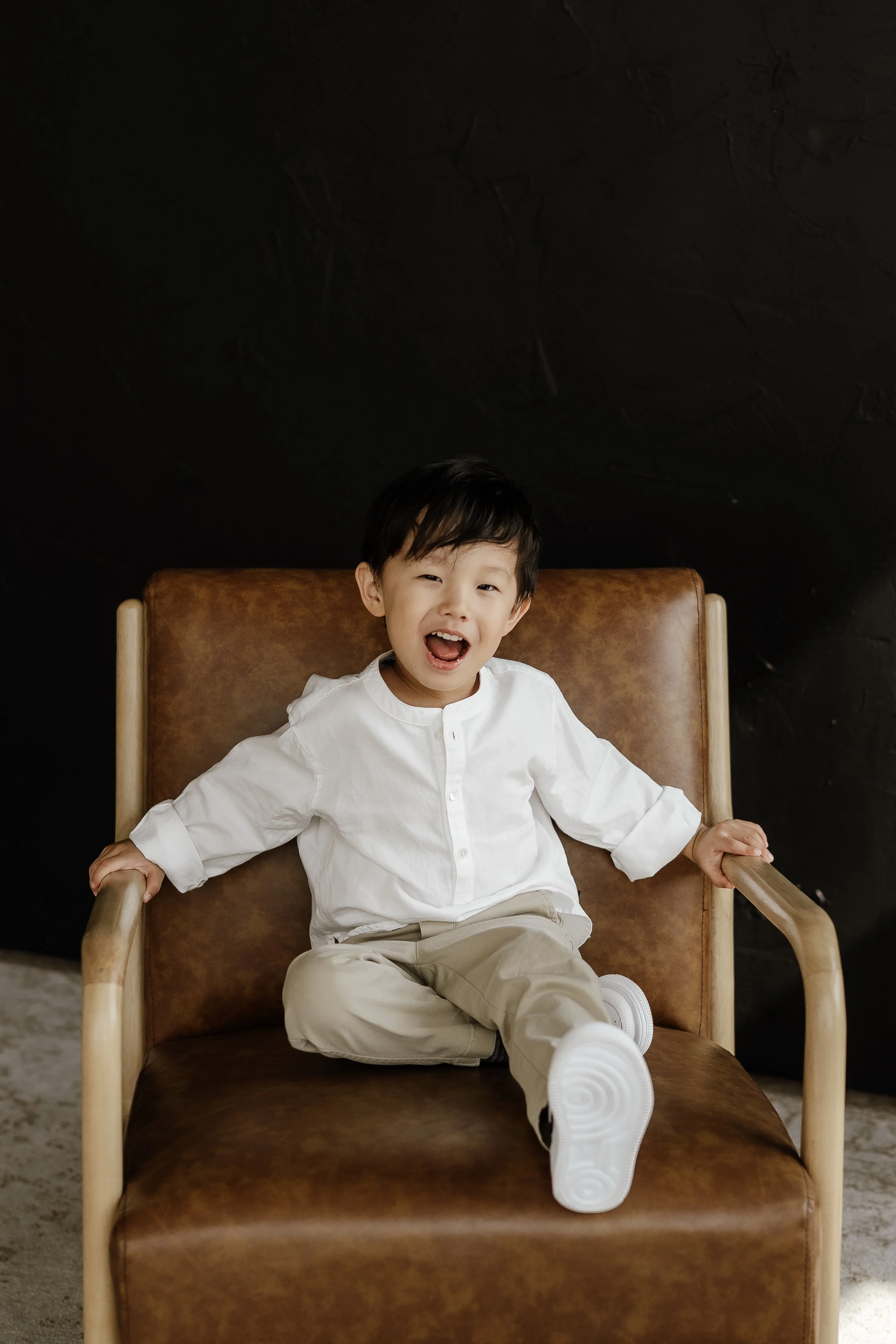 A young boy with black hair smiling while sitting on a brown chair with a black background.