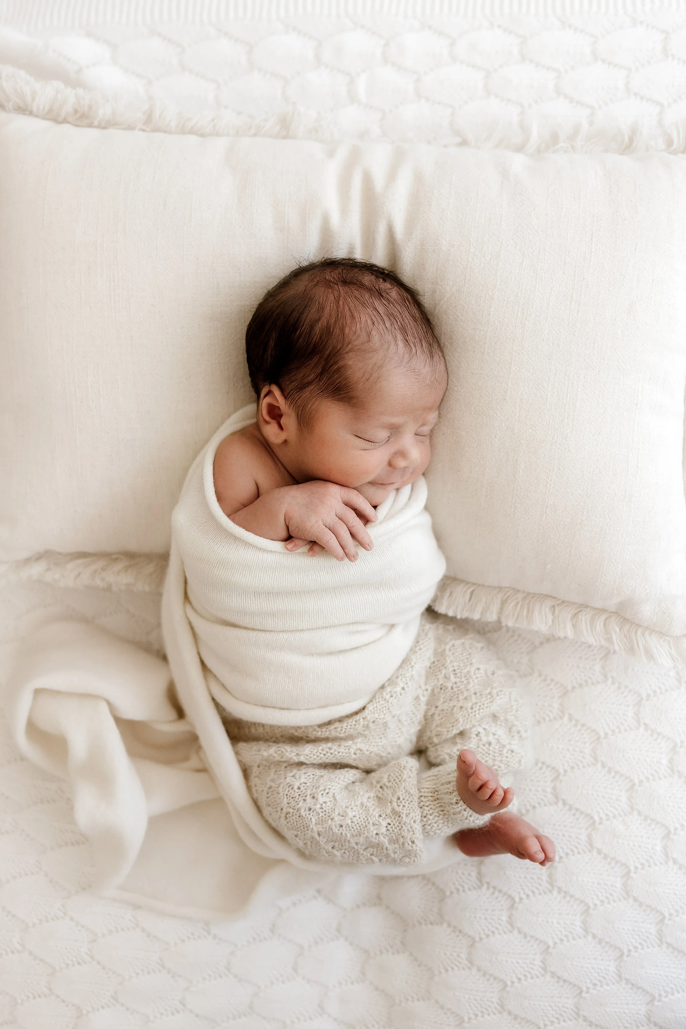 A sleeping newborn baby wrapped in a white blanket, lying on a cream-colored bed with textured sheets and pillows.