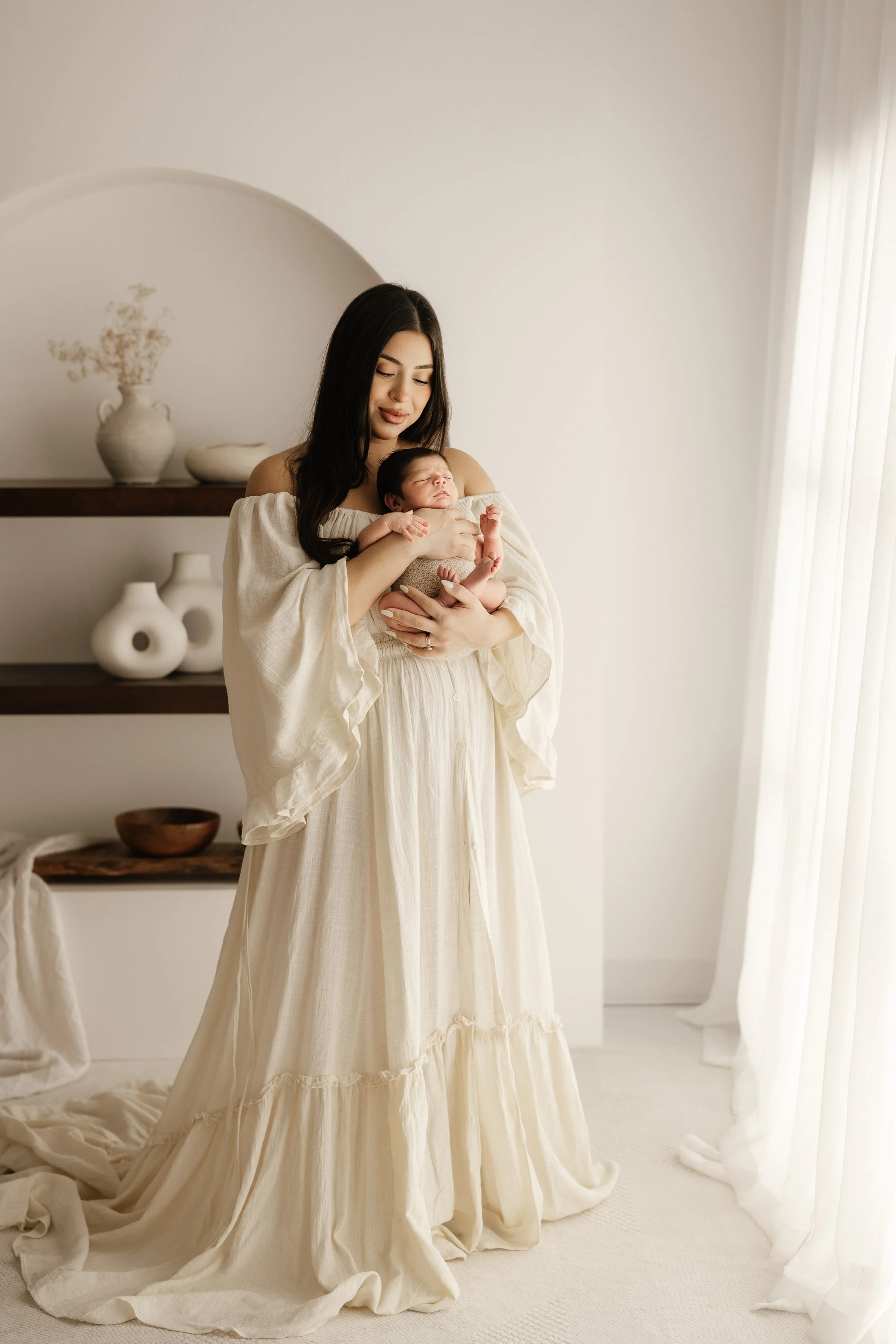 A woman holding a newborn baby indoors near a window with white curtains, dressed in a flowing cream-colored dress, with minimalist decor including vases and bowls on shelves behind her.