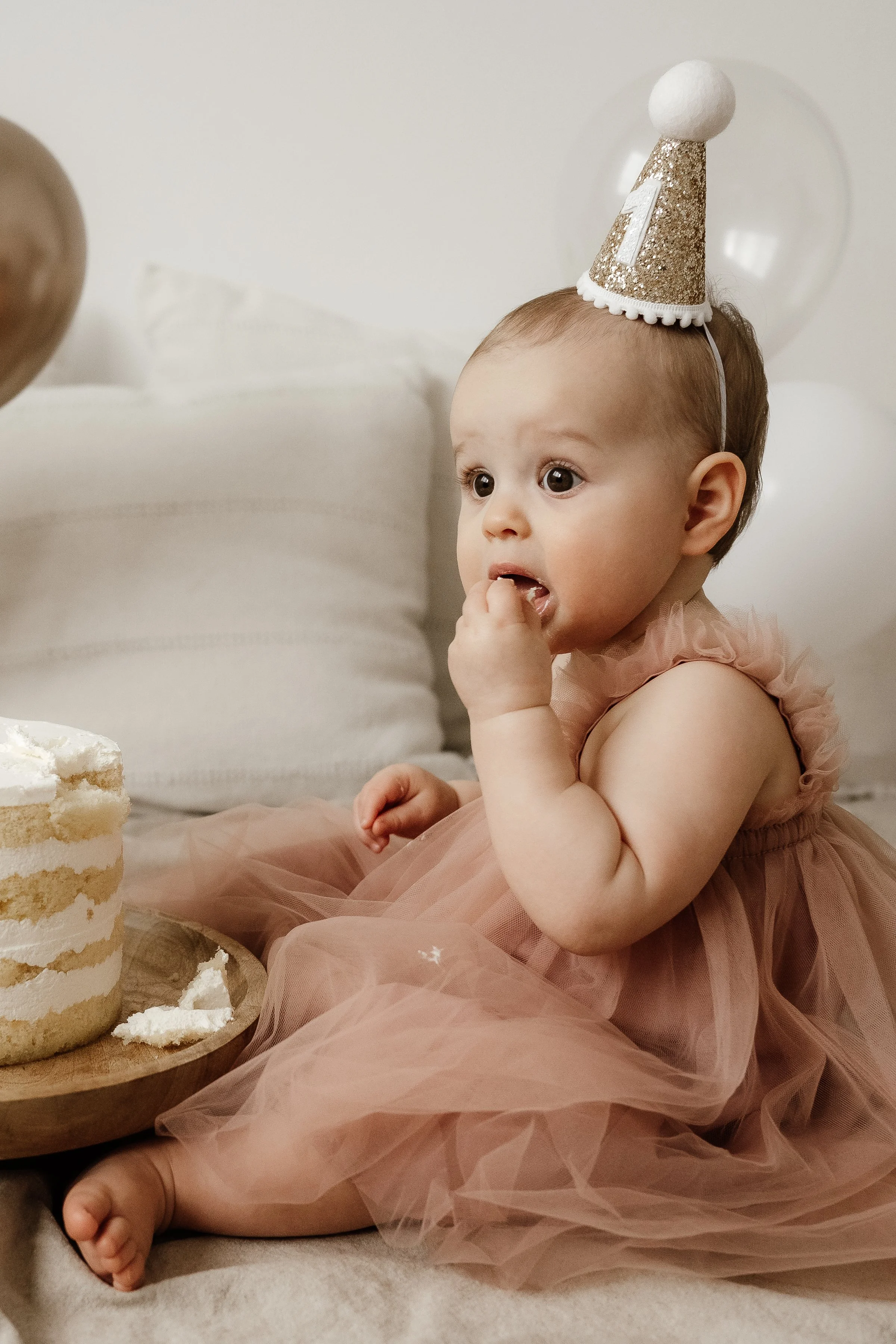 A young child wearing a pink tulle dress and a birthday hat with a number one, sitting on the floor next to a layered birthday cake, with some cake on a wooden plate and her fingers in her mouth, celebrating her first birthday.