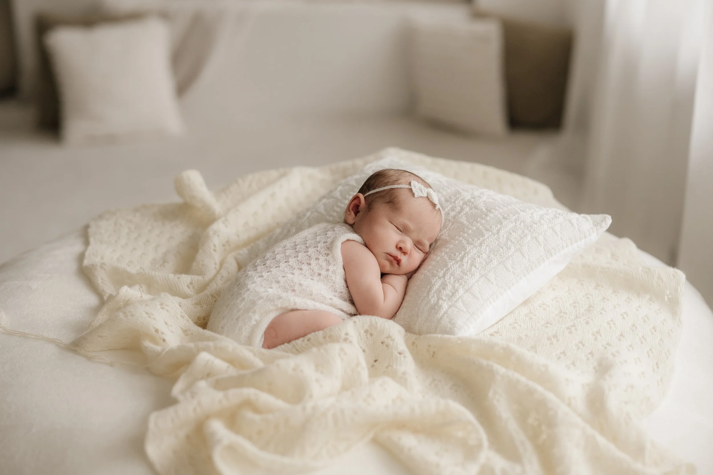 A newborn baby sleeping peacefully on a cream-colored bed with soft linens, wearing a lace outfit and a delicate headband with a bow.