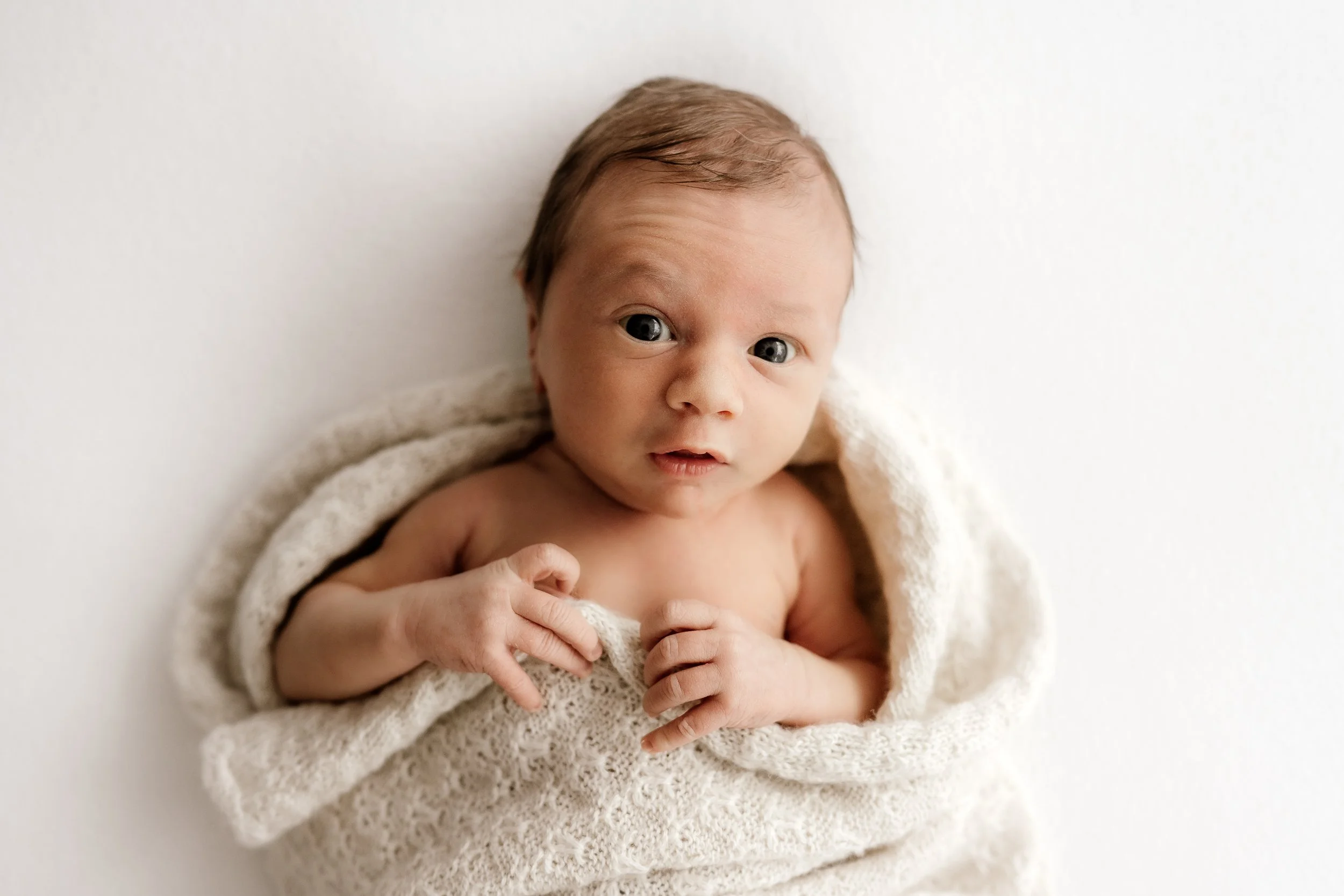 Close-up of a newborn baby with blue eyes, wrapped in a knitted cream-colored blanket, lying on a white surface, looking at the camera.