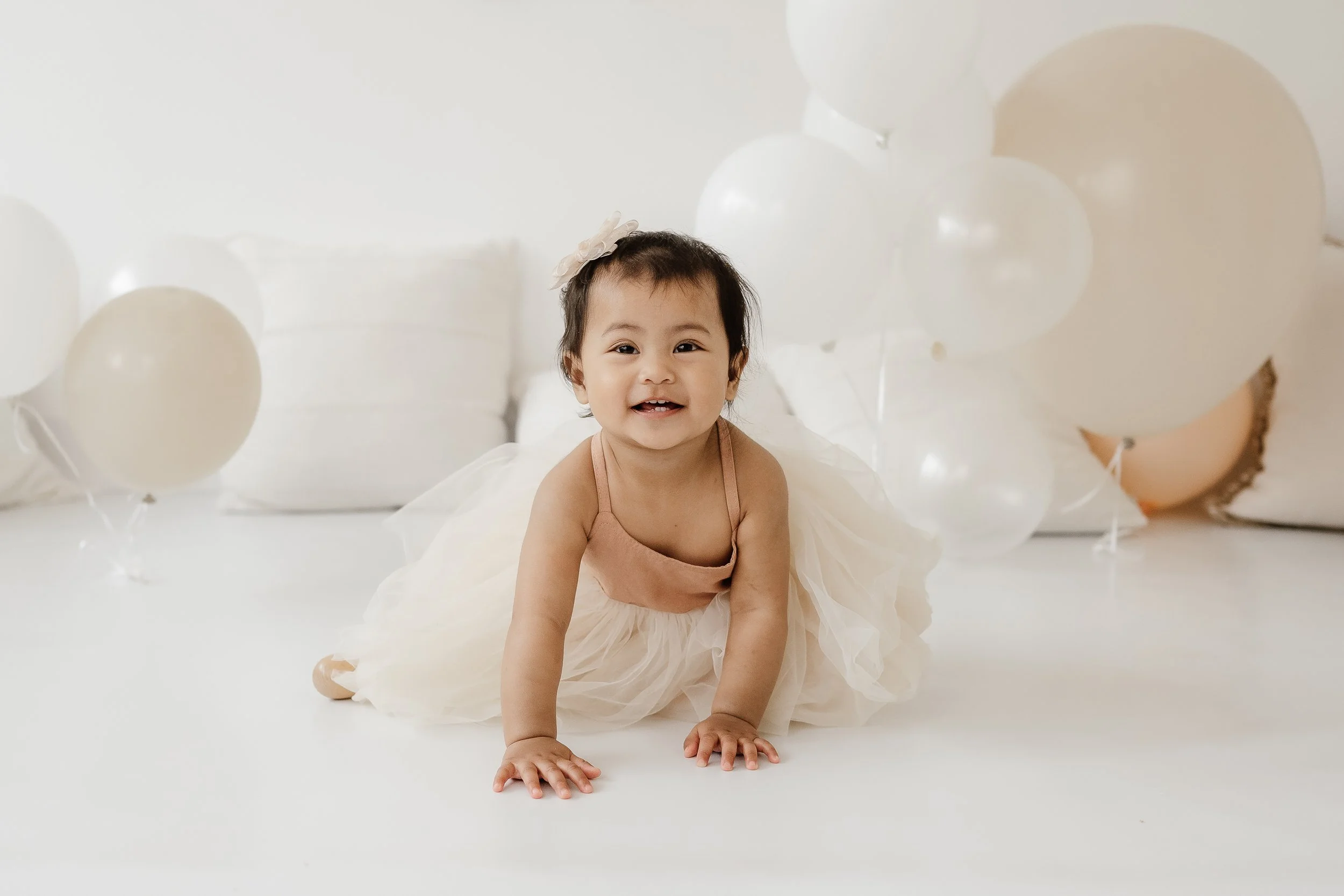 A smiling toddler girl in a beige dress crawling on a white floor surrounded by white balloons in a bright, neutral-toned room.