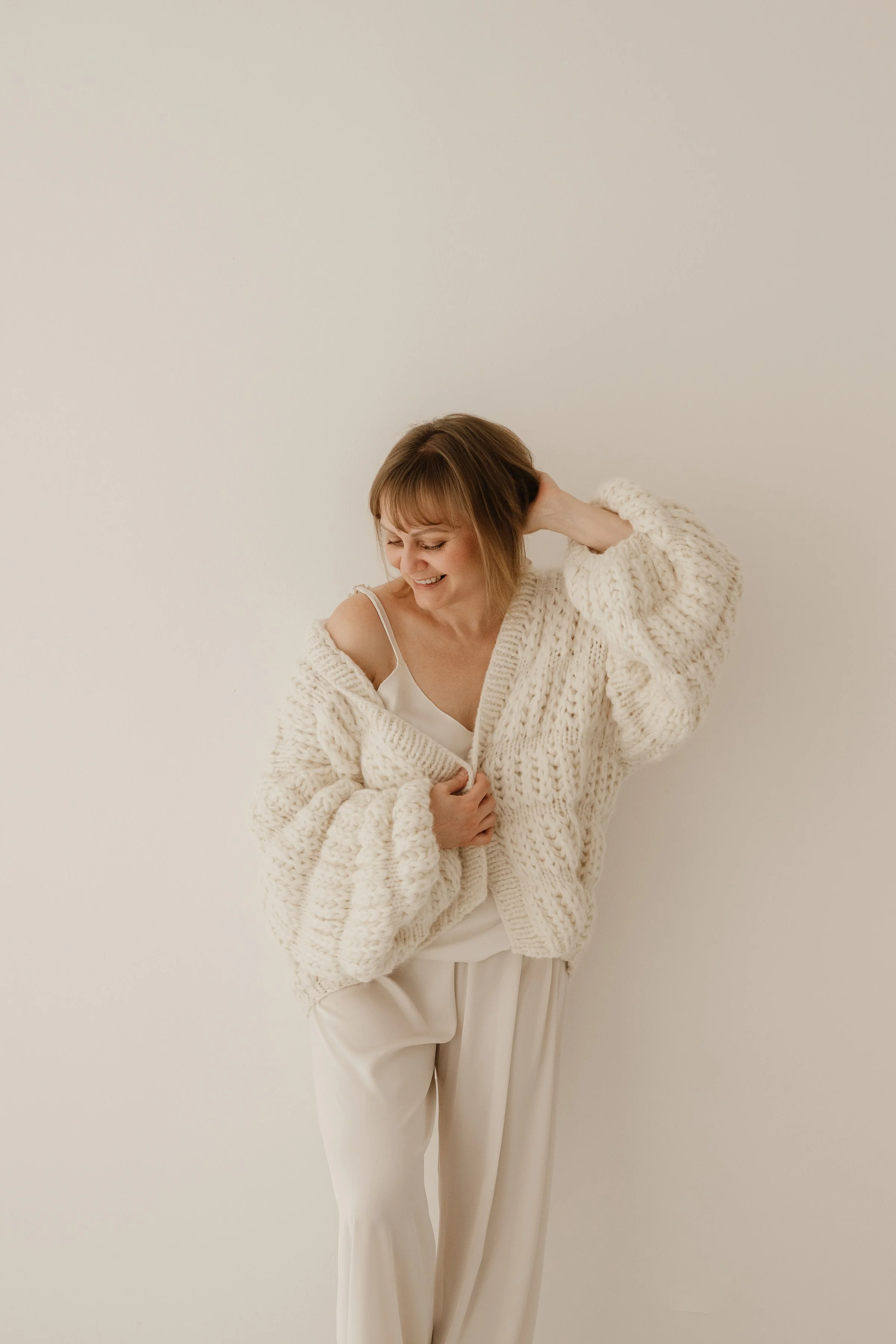 A woman in a cozy, chunky knit cream sweater and white outfit smiling and laughing while touching her hair against a plain, light-colored wall.