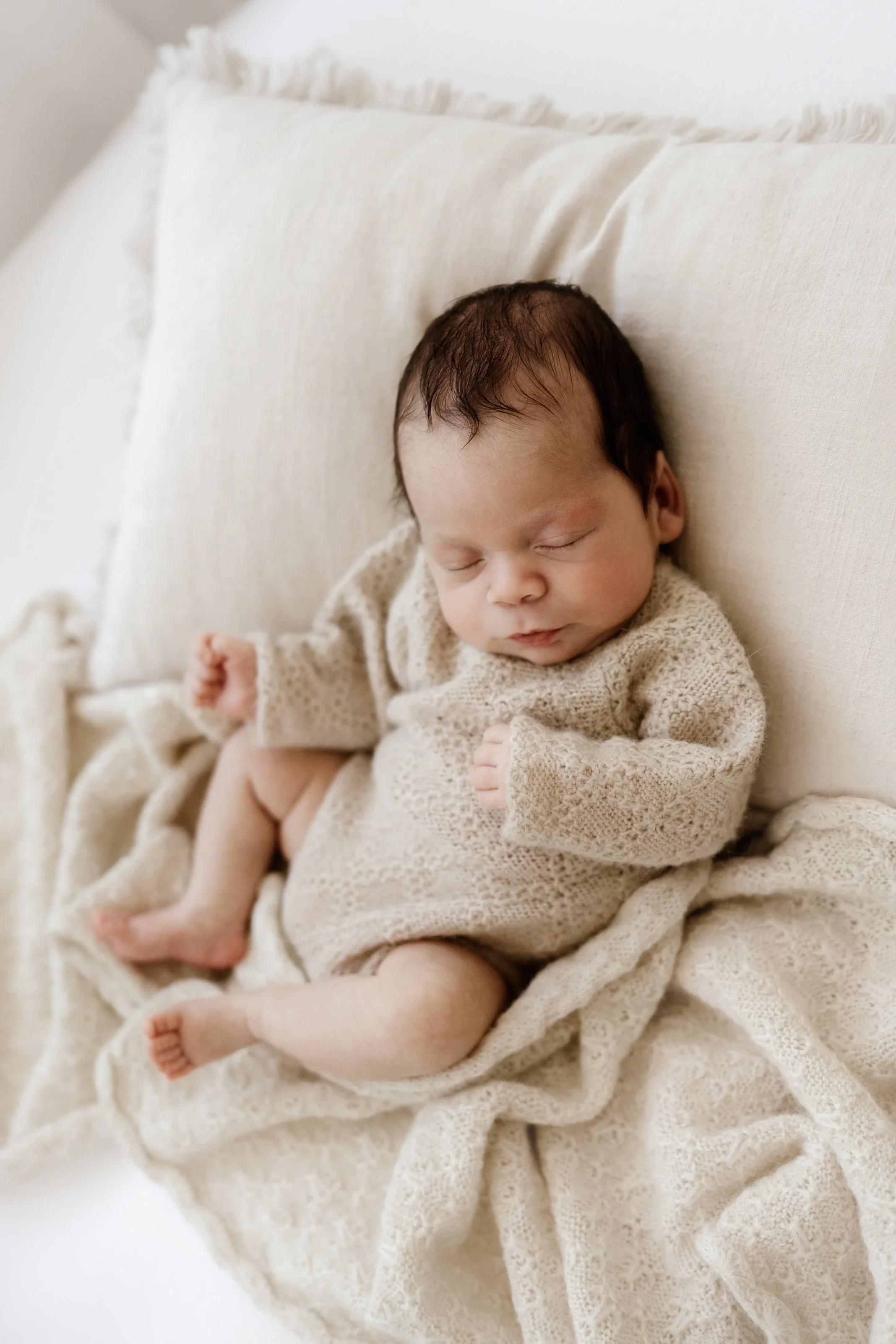 Precious newborn baby sleeping on cream-colored bedding, dressed in a cozy beige knitted outfit.
