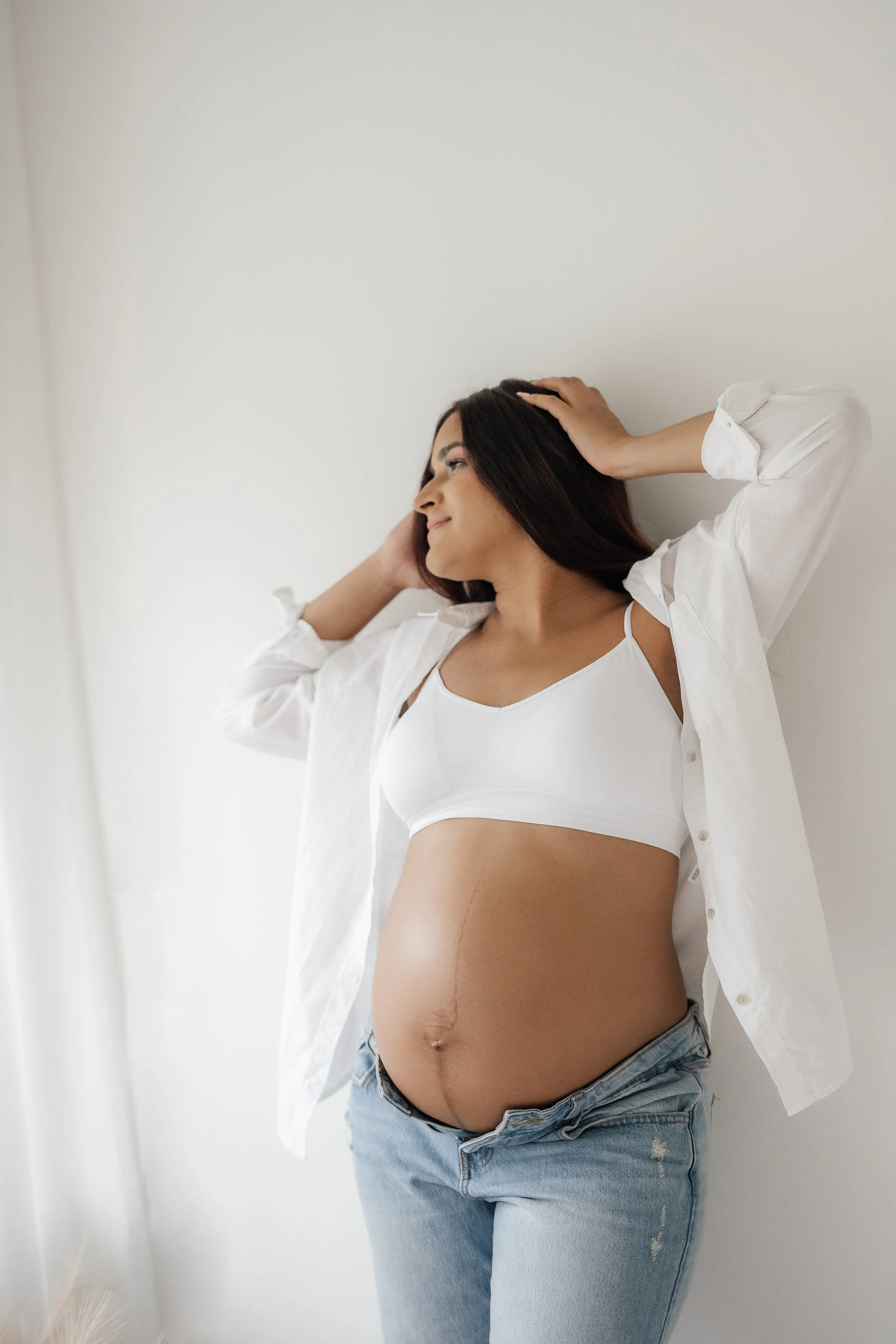 A pregnant woman with long dark hair wearing a white tank top and an open white shirt, standing against a white wall, smiling and posing with her hands in her hair.