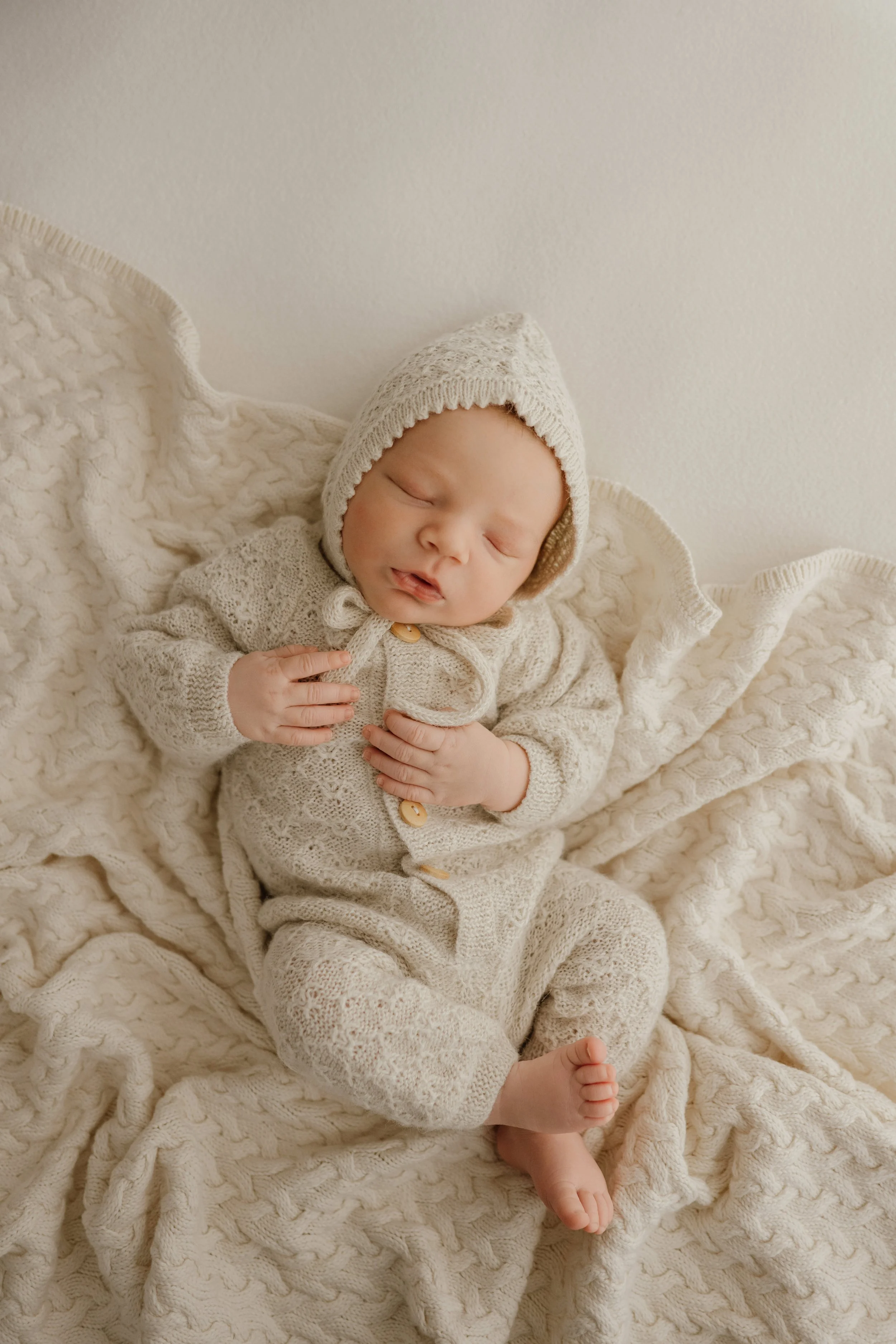 A sleeping baby dressed in a cream-colored knitted outfit and bonnet, lying on a cozy cable-knit blanket.