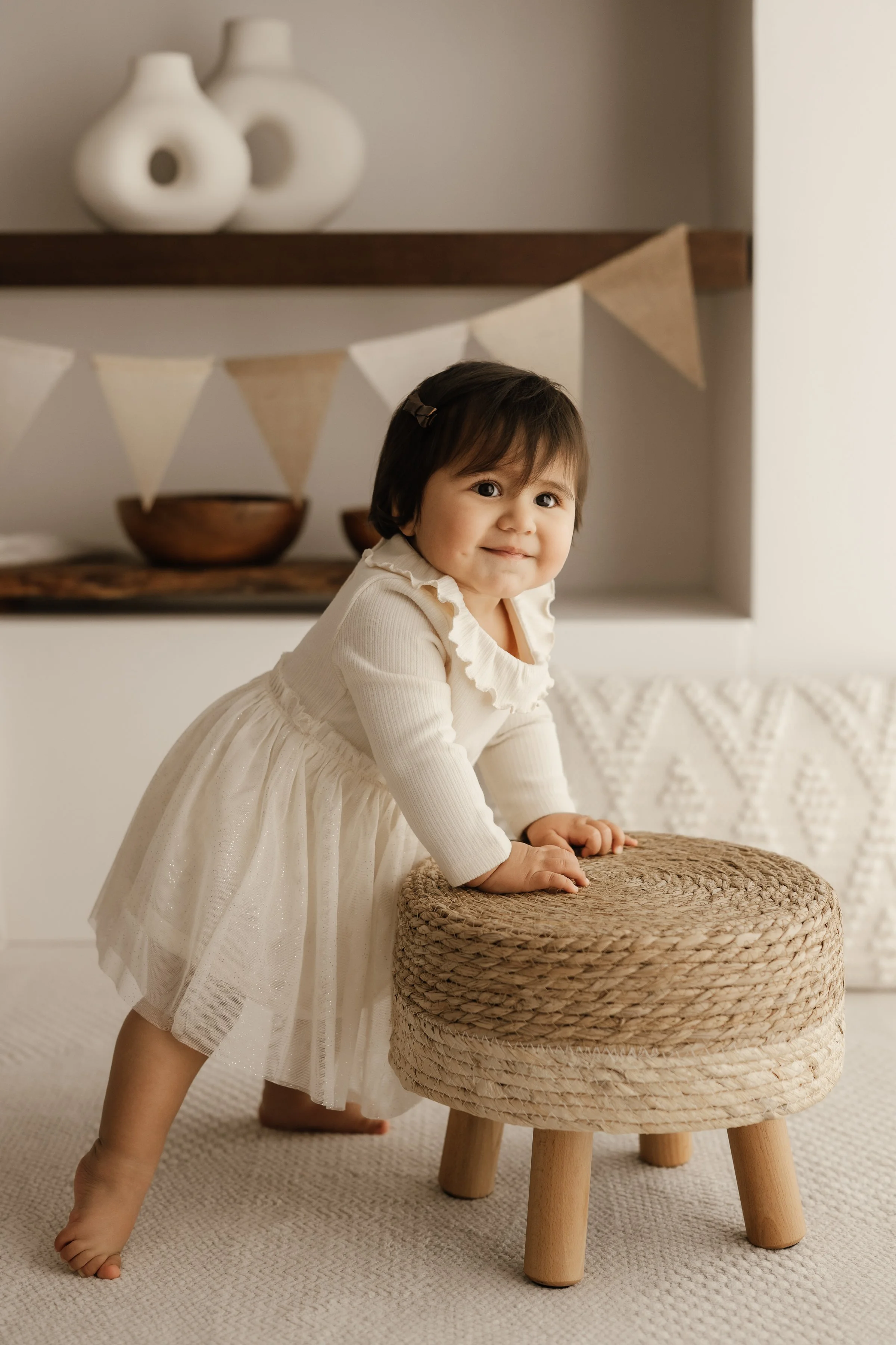 Young girl in a white dress playing with a small round woven ottoman in a cozy room with neutral decor.