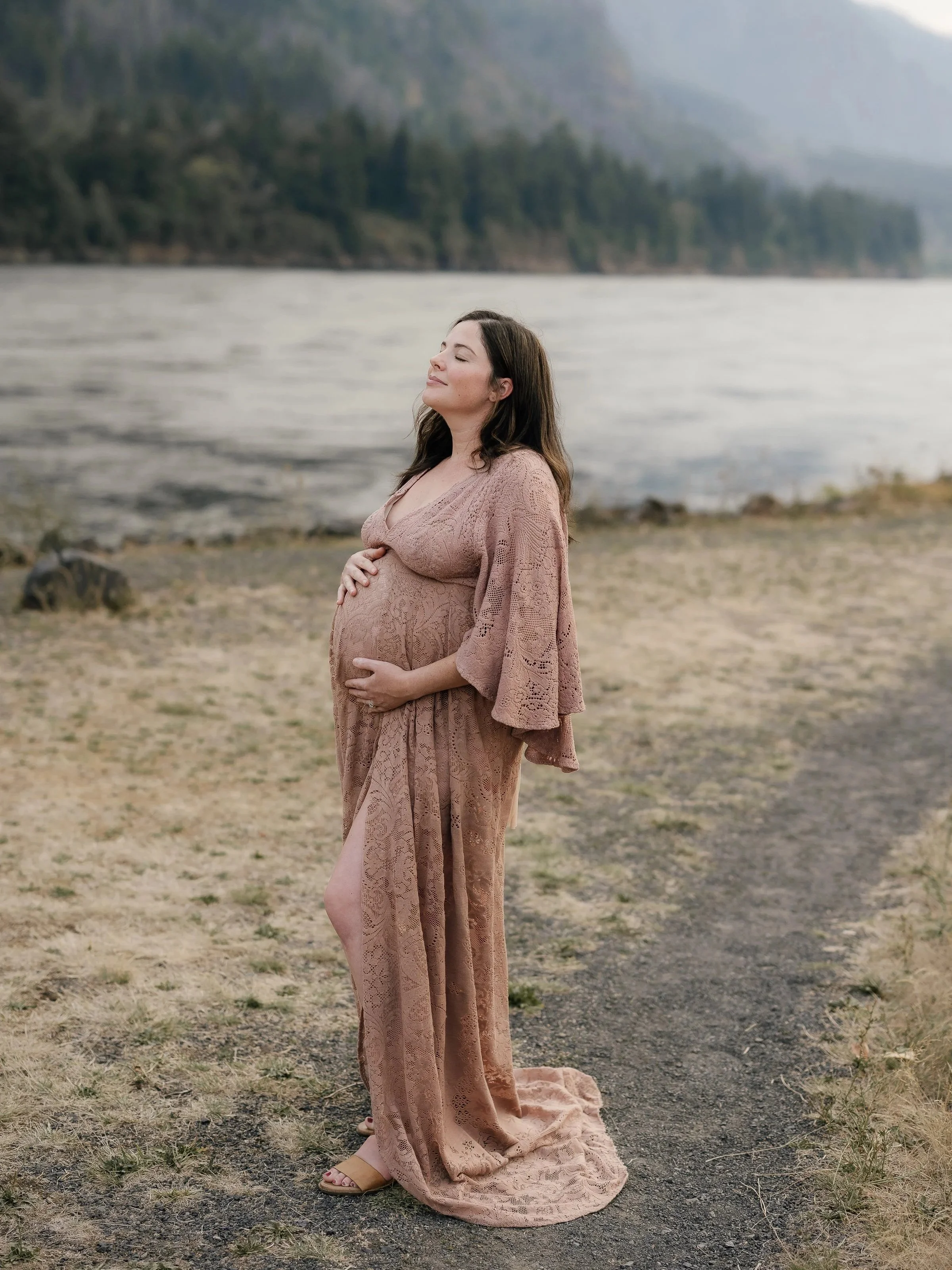 Pregnant woman in a pink lace dress standing on a natural path near a body of water with mountains and trees in the background, tending to her belly with a peaceful expression.