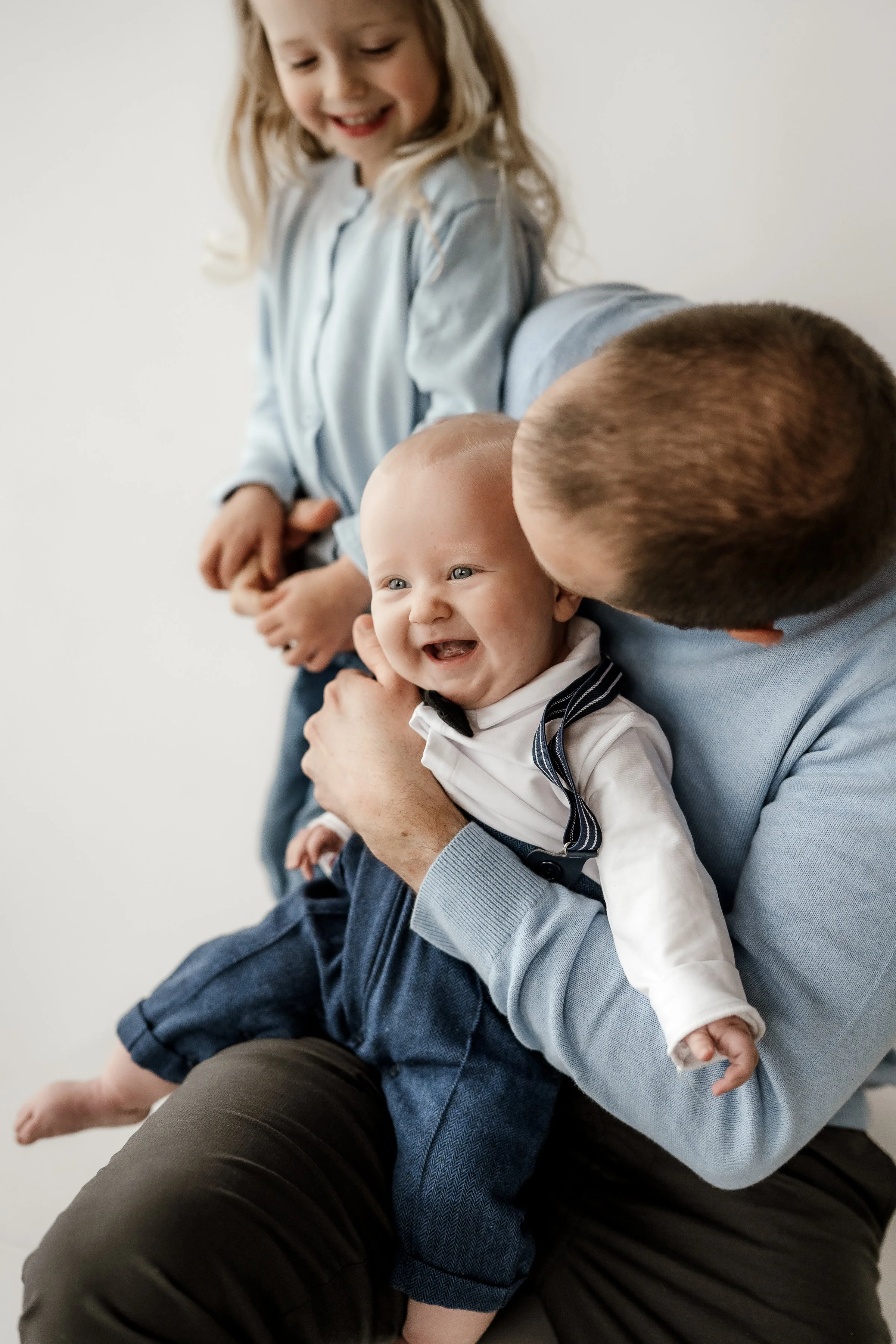 A man holds a smiling baby boy dressed in a tuxedo, with a woman and a girl in the background, all enjoying a joyful moment together.