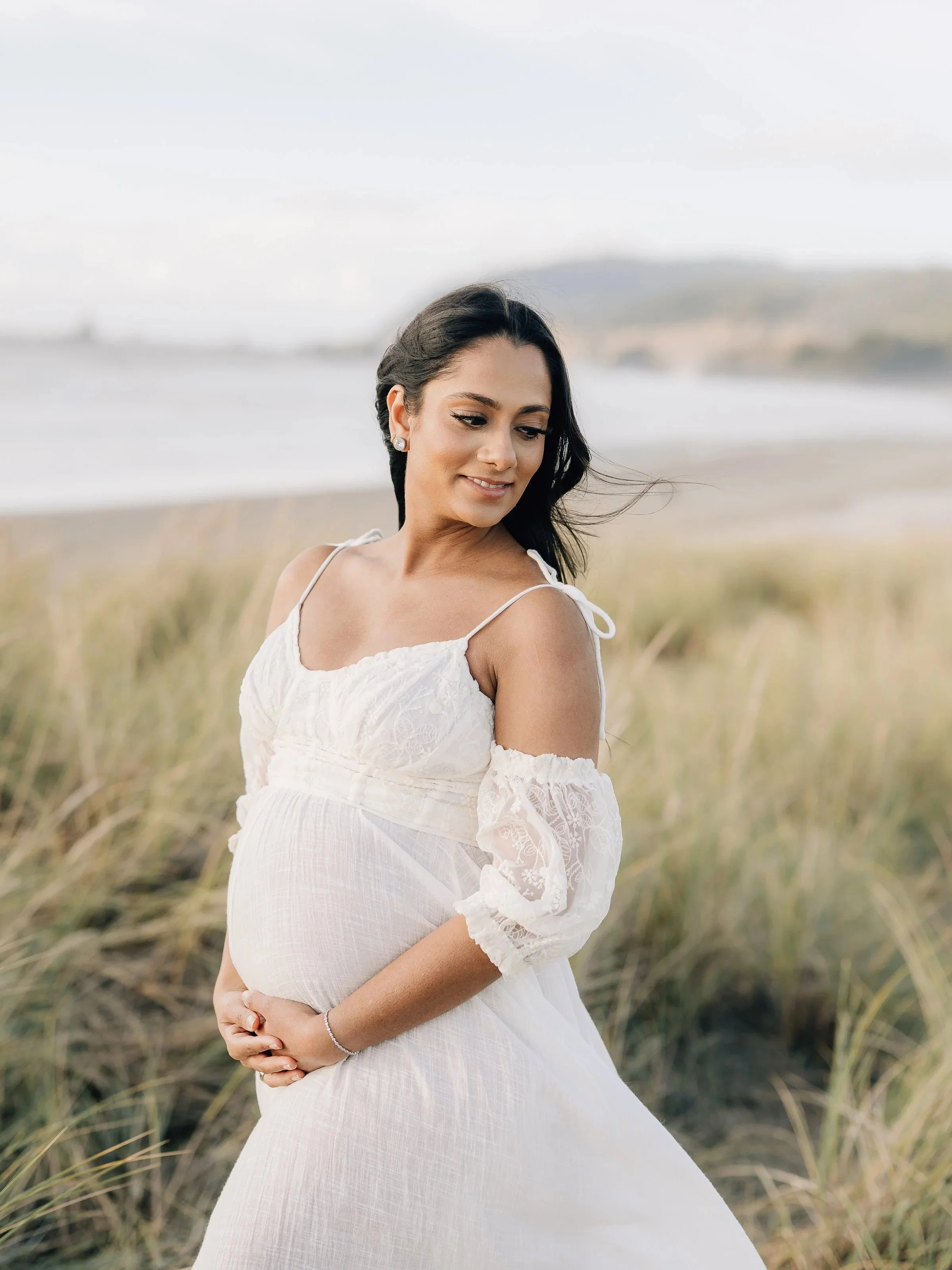 A pregnant woman with dark hair in a white lace dress standing outdoors on a beach, holding her belly, with grasses and the ocean in the background.