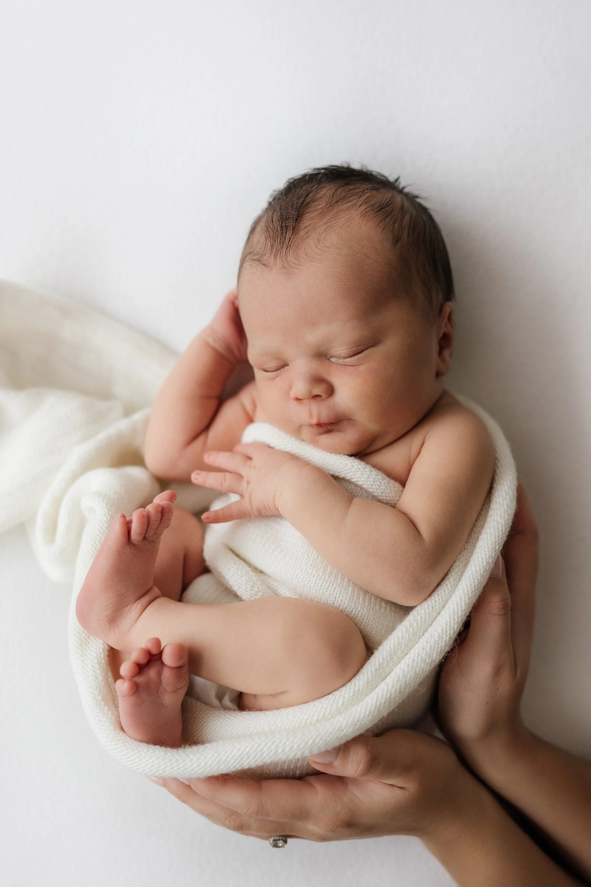 A newborn baby sleeping peacefully, wrapped in a white blanket, with one hand touching its face and the other resting on its chest, held gently by an adult's hands against a white background.
