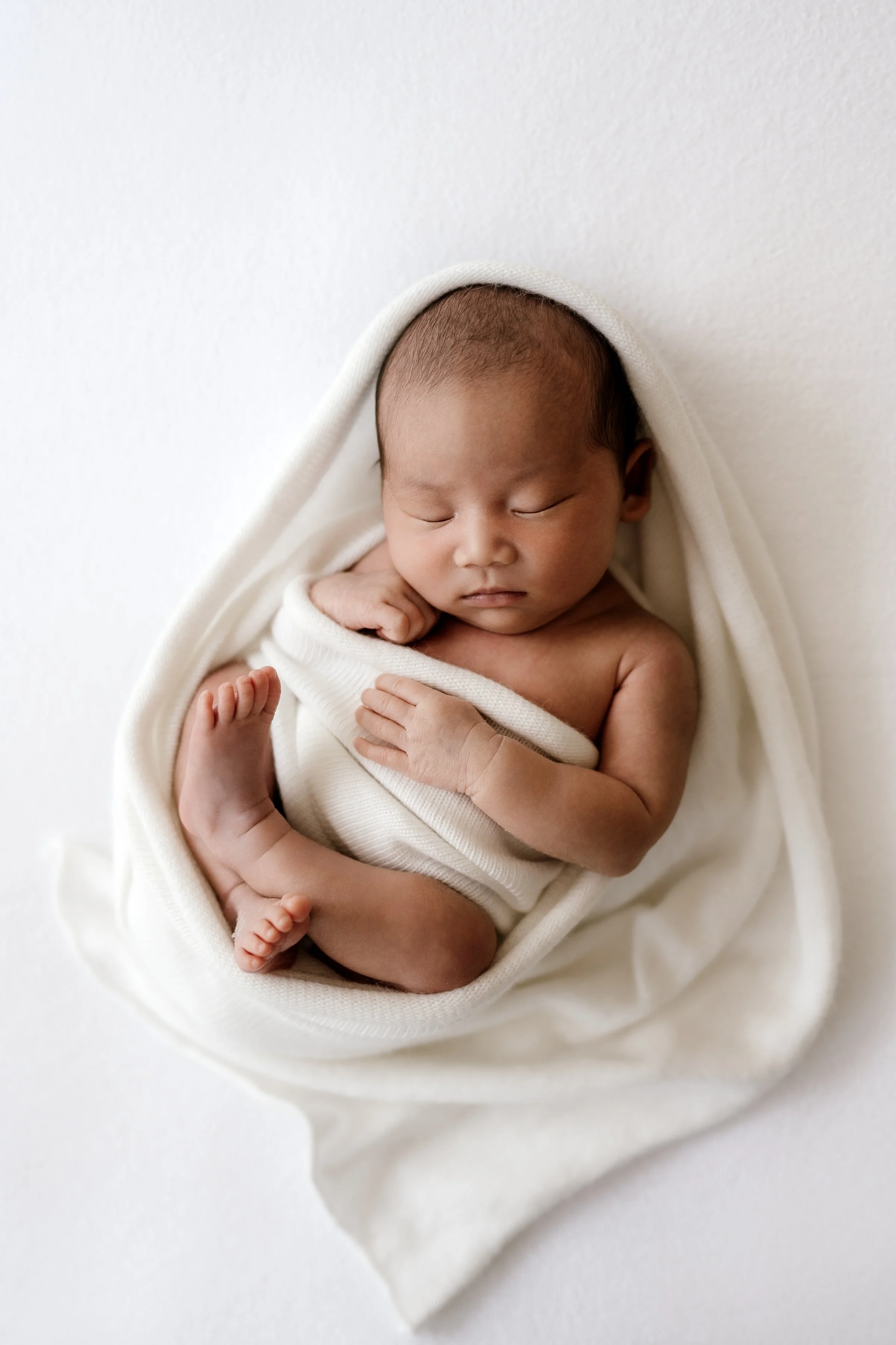 Newborn baby peacefully sleeping, wrapped in a cream-colored blanket, lying on a white surface.