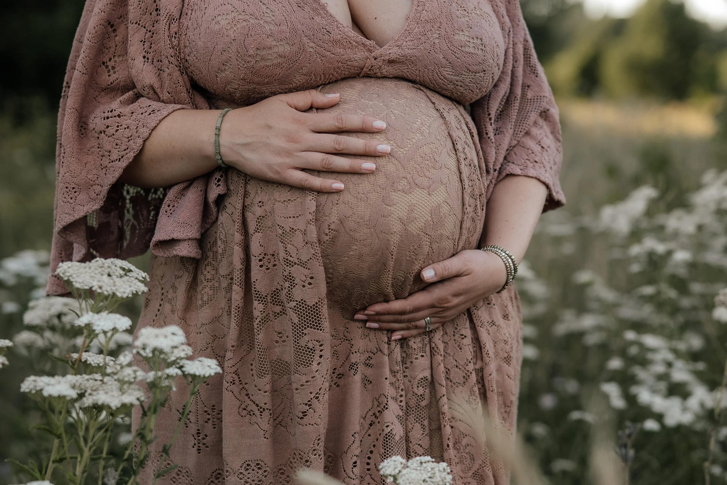 A pregnant woman wearing a pink lace dress and accessories standing in a field of white flowers, gently holding her belly with both hands.