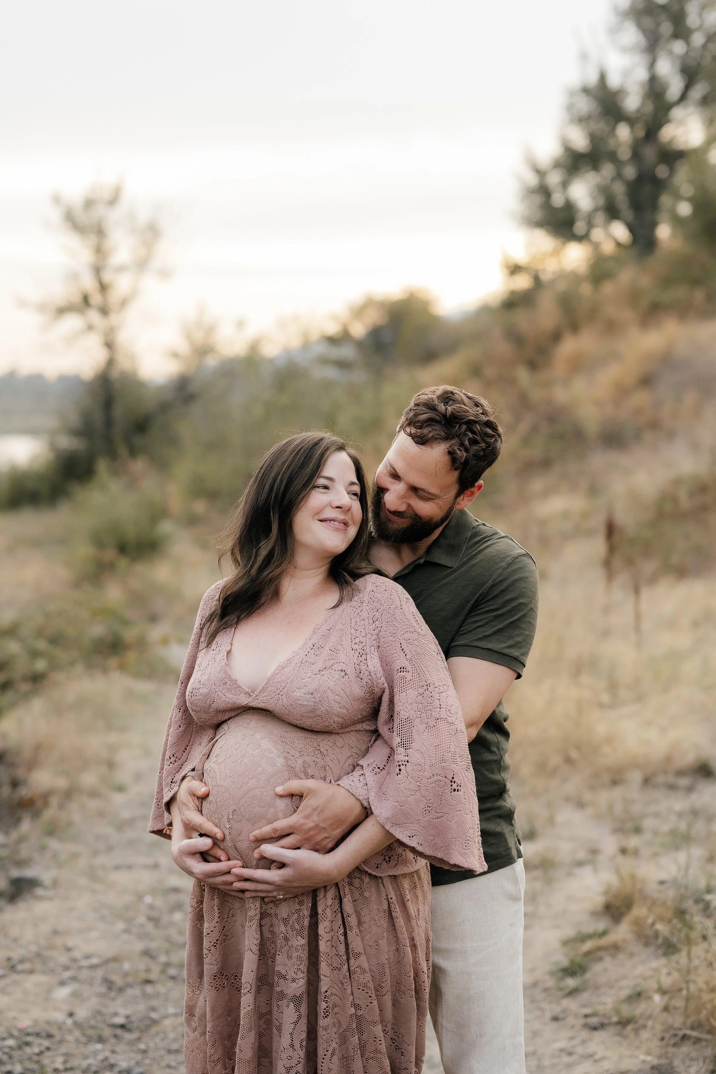 A pregnant woman and her partner smiling and holding her baby bump outdoors in a natural setting with trees and a river in the background during sunset.