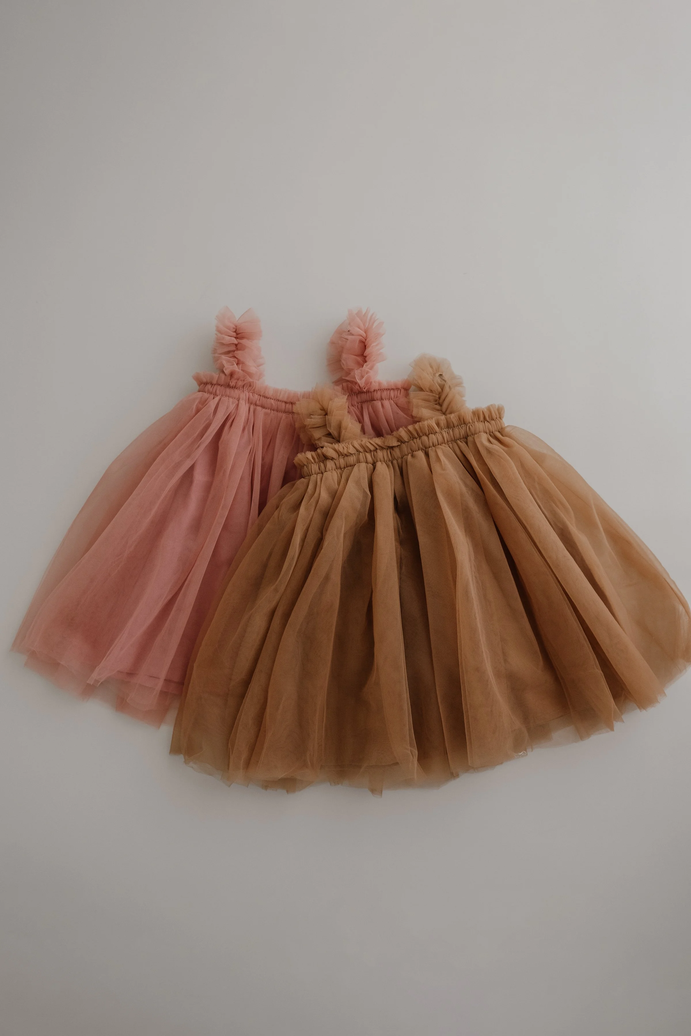 Two toddler tutu skirts, one pink and one beige, with ruffled straps, lying on a white surface.