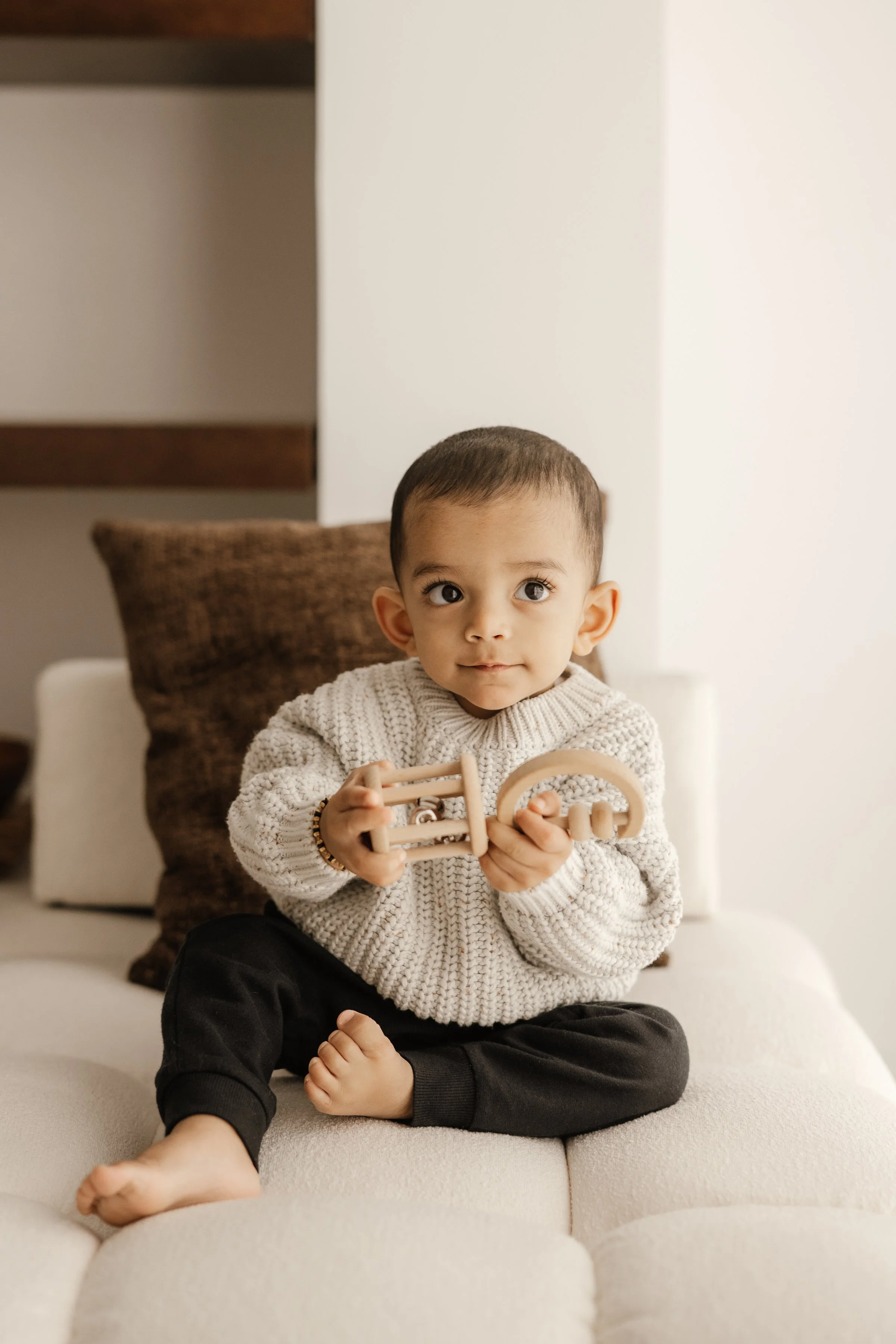A young child with short dark hair, wearing a chunky beige sweater and black pants, sits cross-legged on a white couch holding a wooden teething toy, with a brown cushion behind him and a modern interior background.