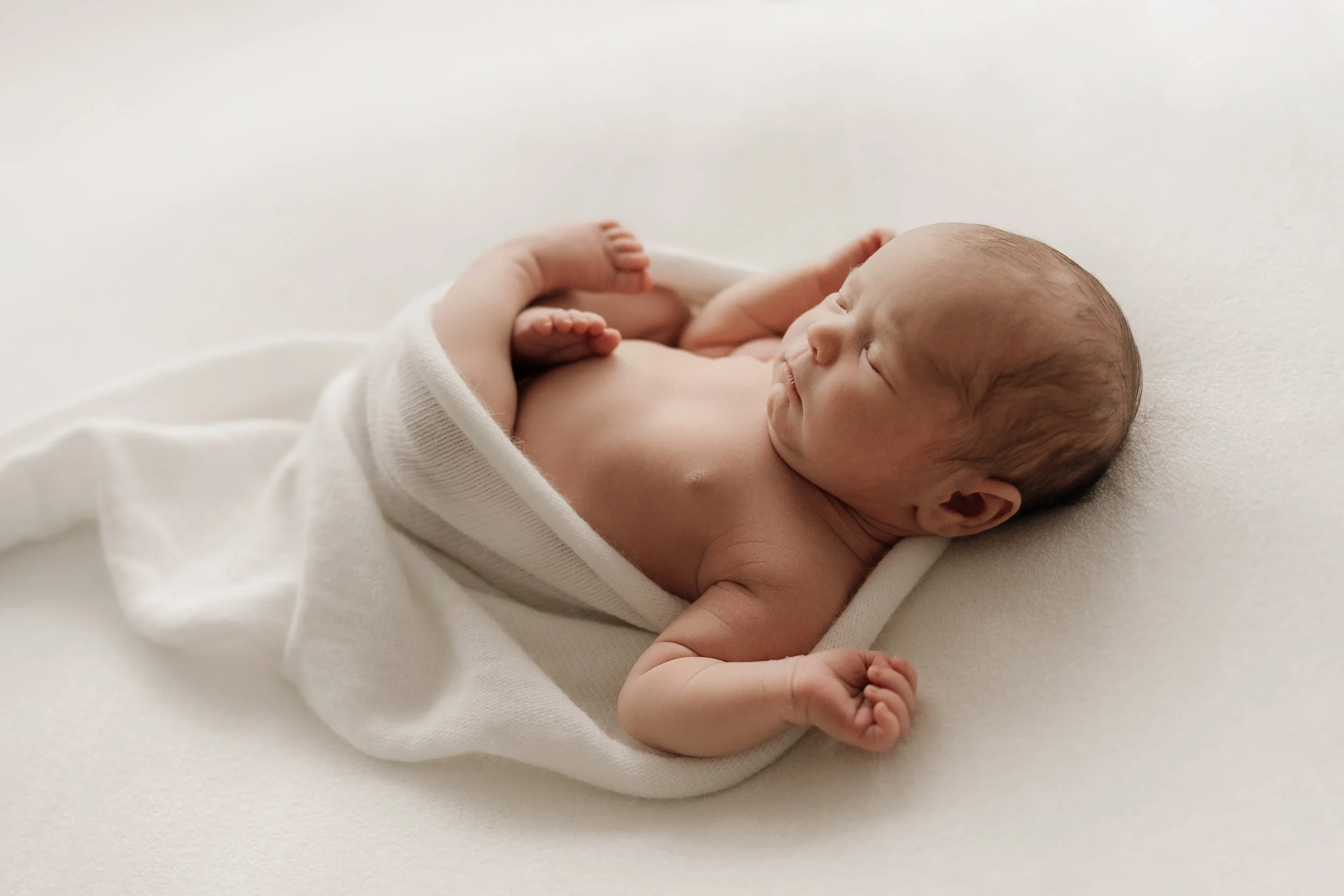 A newborn baby lying on a white surface, partially wrapped in a white cloth, with eyes closed.