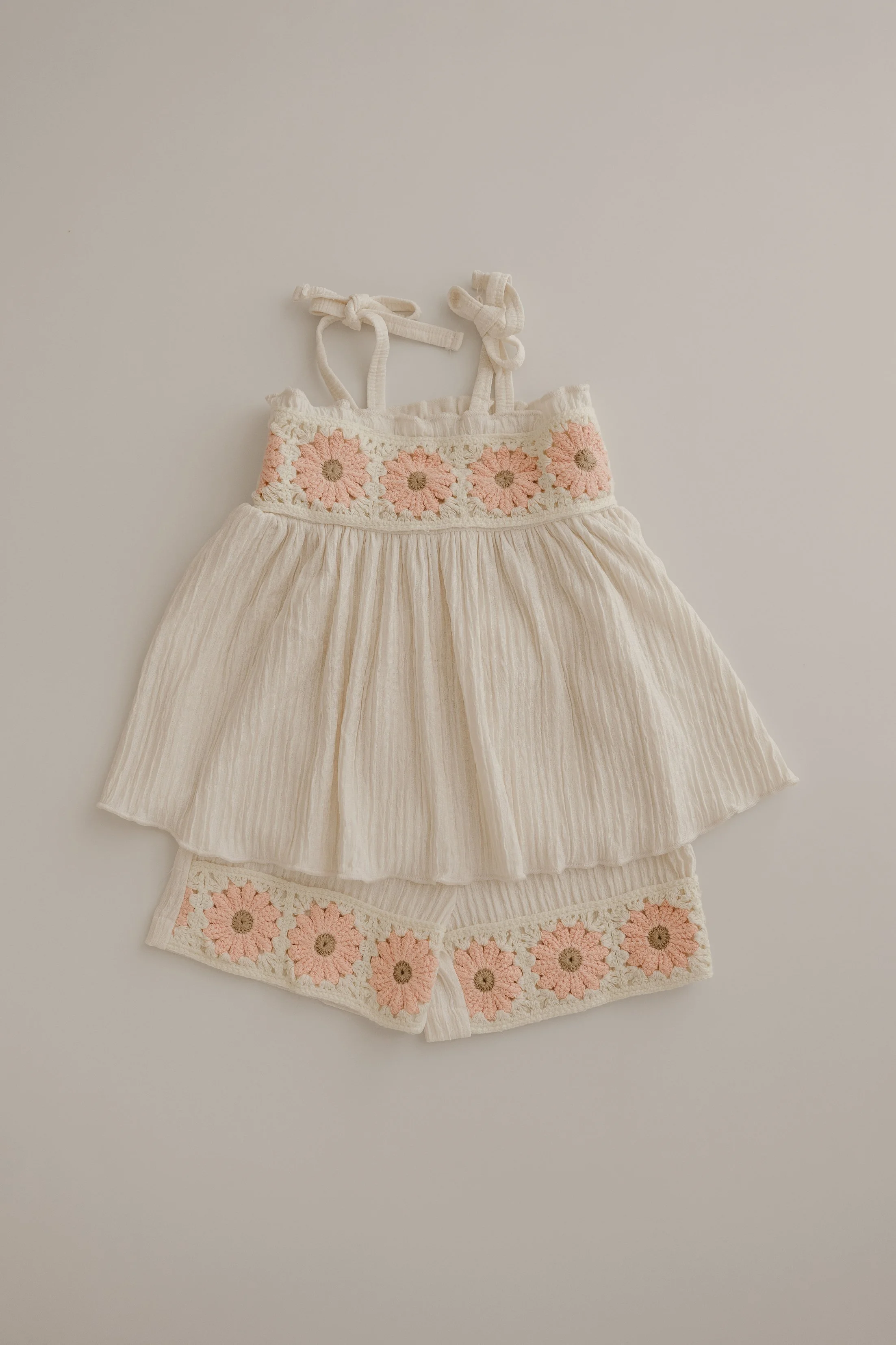 A matching set of children's clothing, including a cream-colored sleeveless top and shorts, both with pink and beige crochet flower embellishments along the waistband and hem.