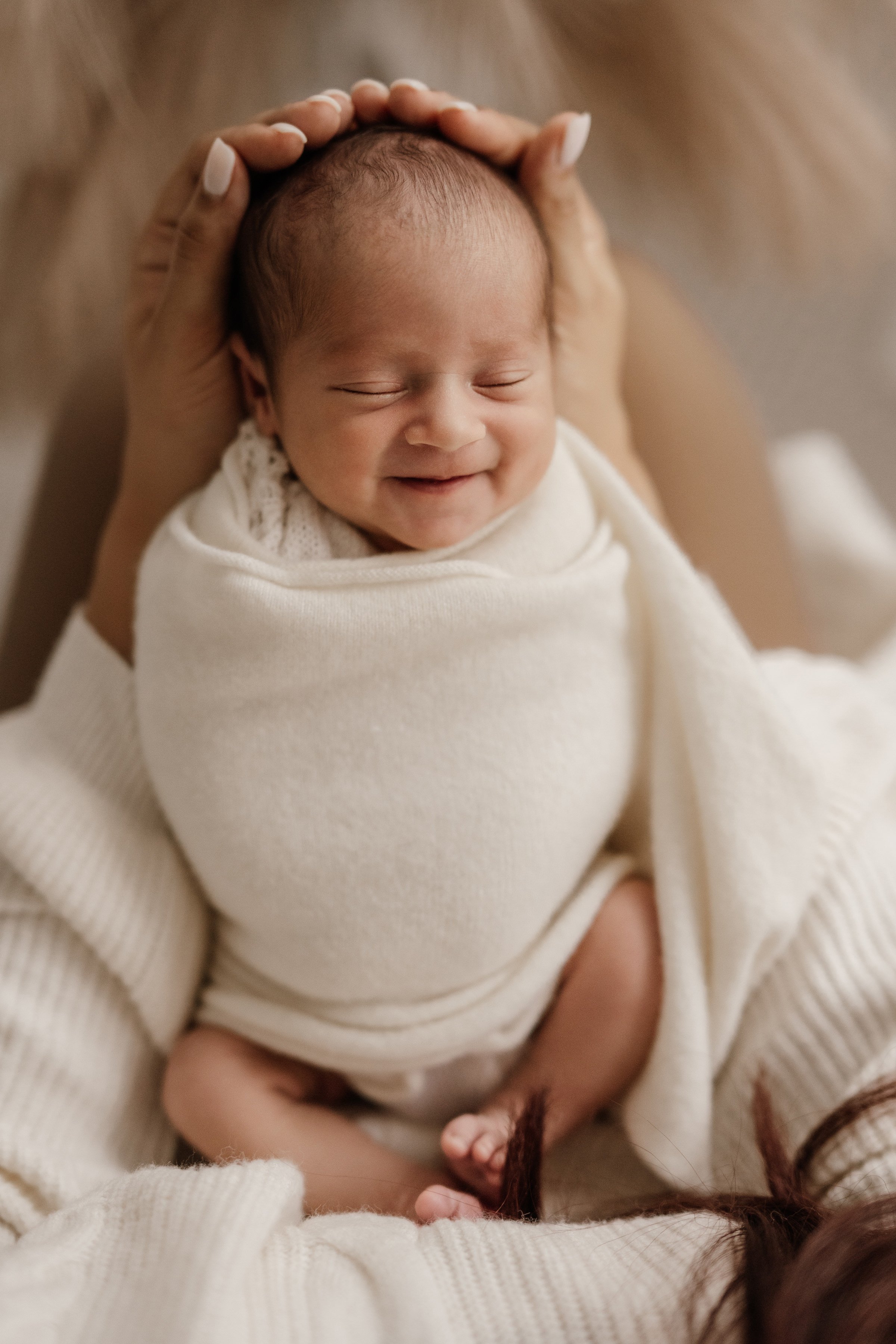 A smiling newborn baby with closed eyes wrapped in a cream-colored blanket, being gently held by an adult