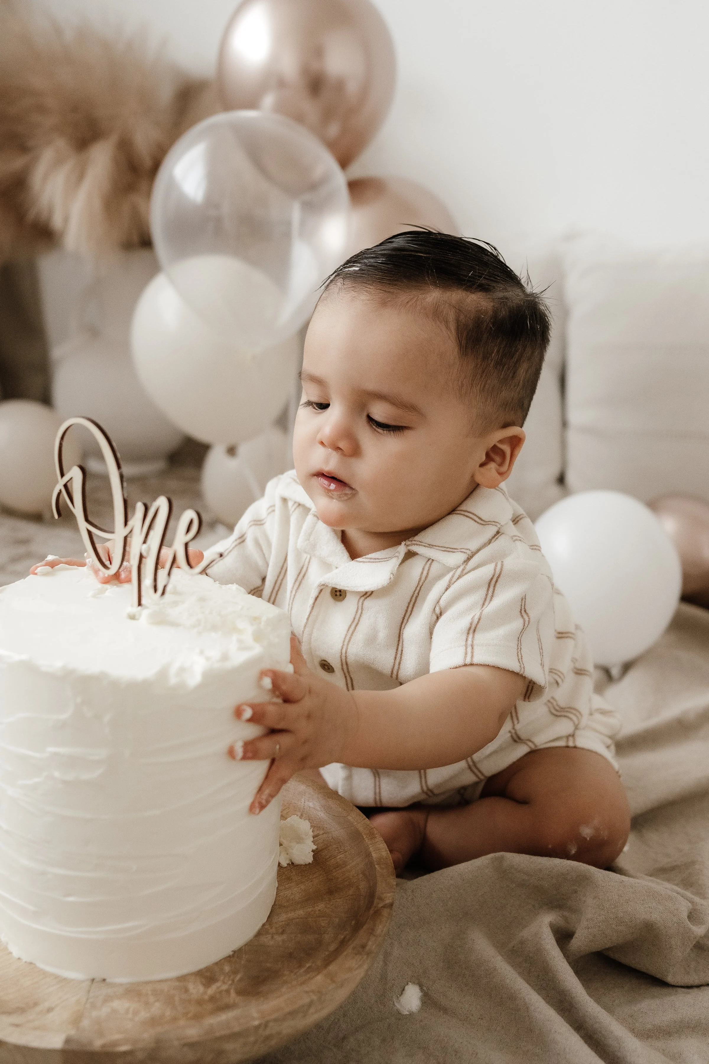 A young boy sitting on the floor, holding a white birthday cake with a 'One' topper, surrounded by white and blush balloons, celebrating his first birthday.