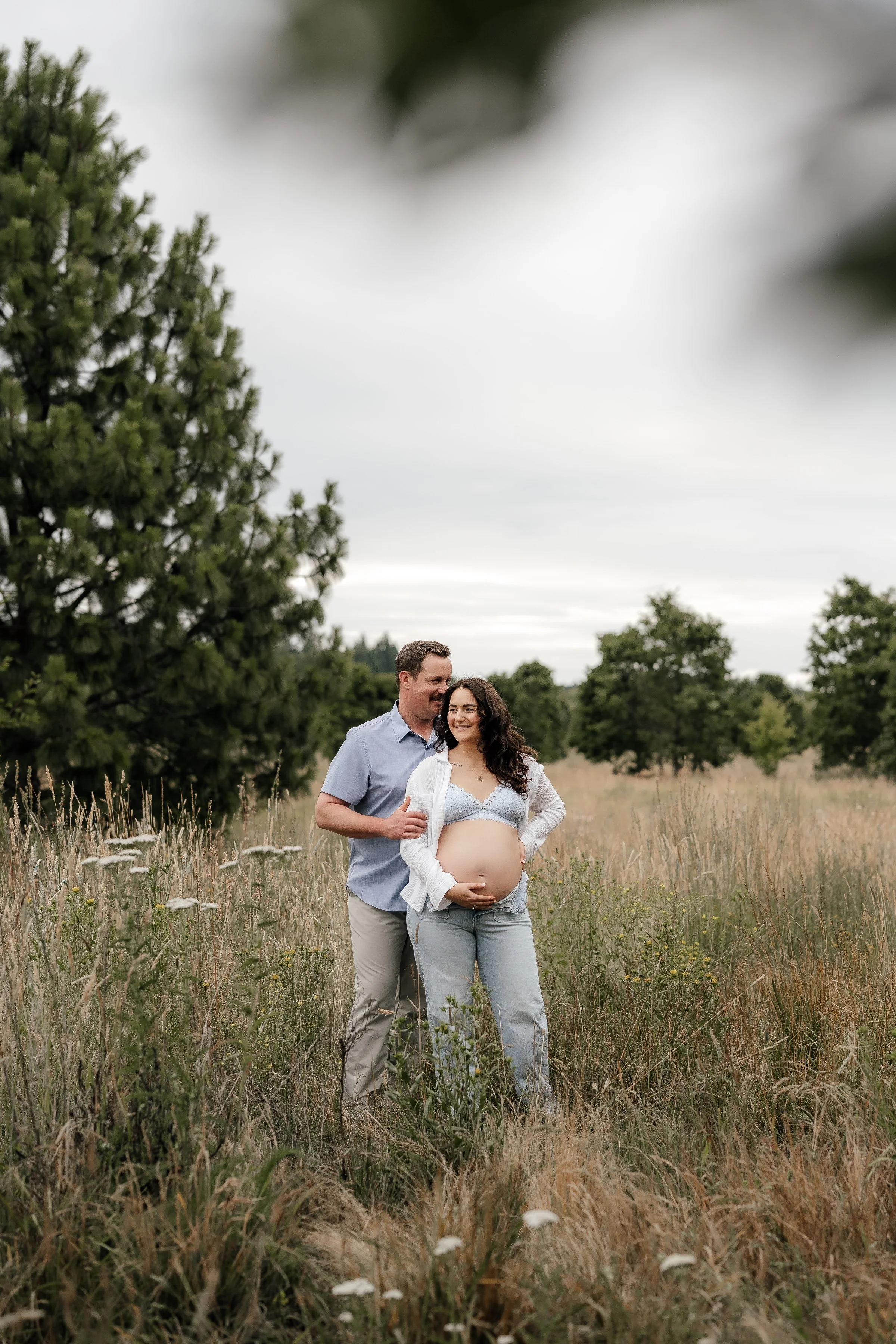 A pregnant woman and a man standing in a field of tall grass and wildflowers, with trees in the background, on a cloudy day.