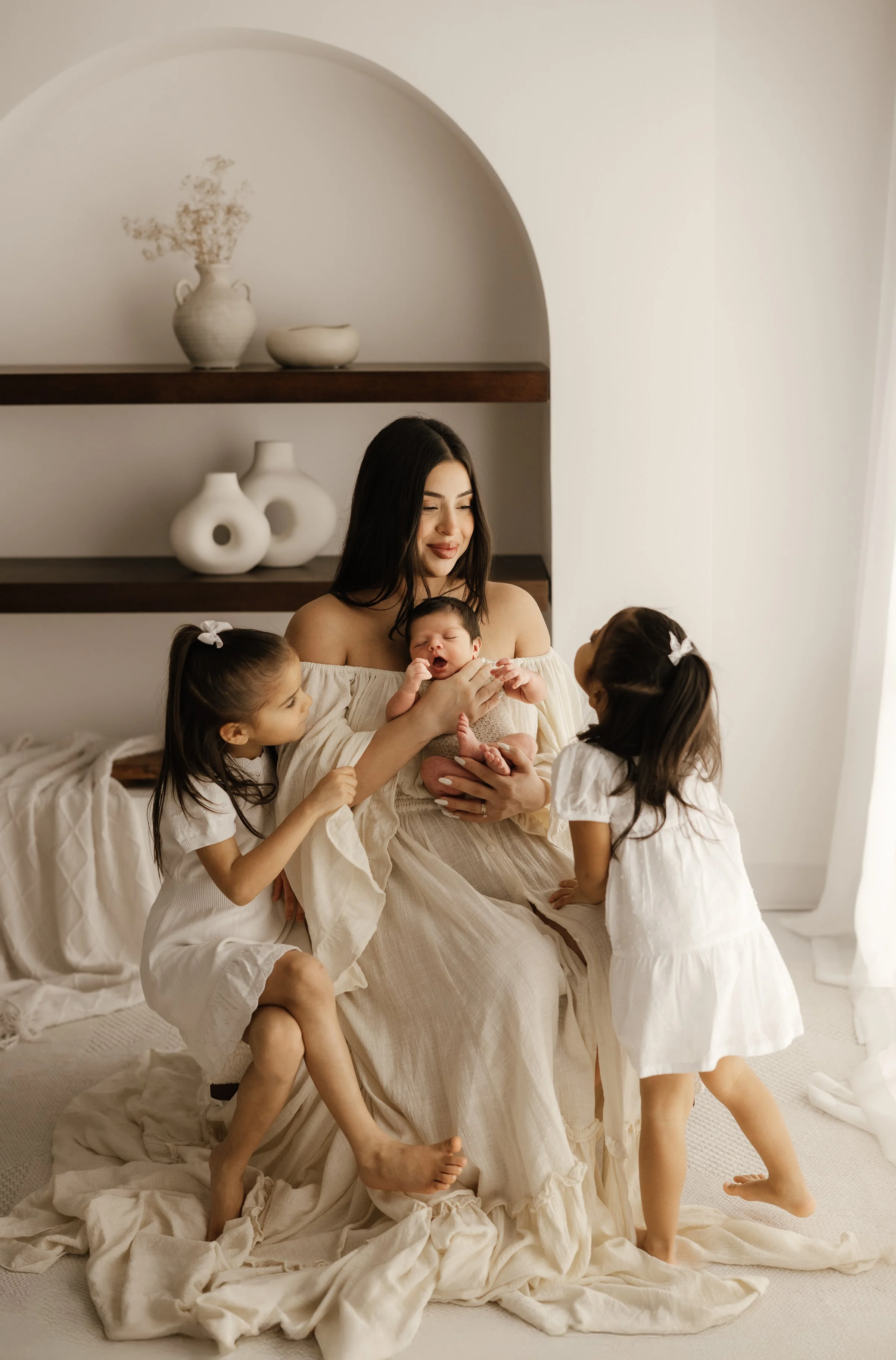 A woman holding a newborn in a beige dress while two young girls in white dresses look at the baby. They are in a softly lit room with white walls and decorative shelves with white vases.