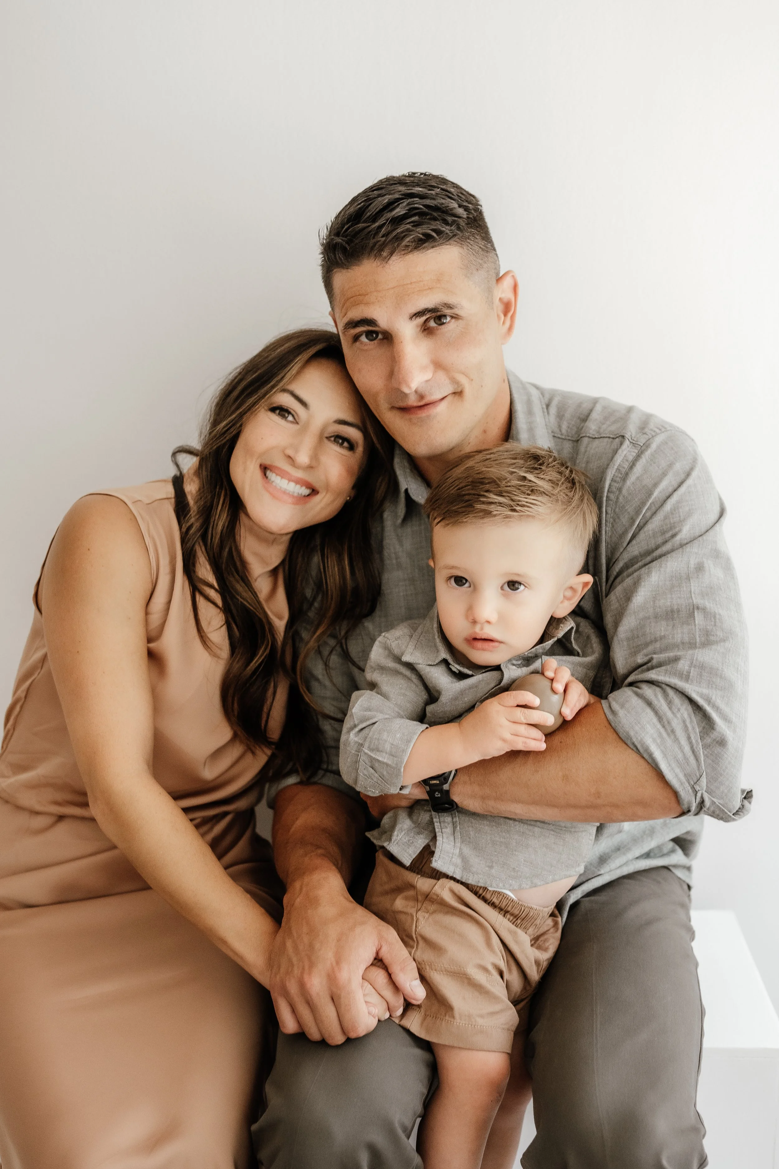A happy family of three sitting together against a plain white background, with a woman with long brown hair, a man with short dark hair, and a young boy with short blond hair.
