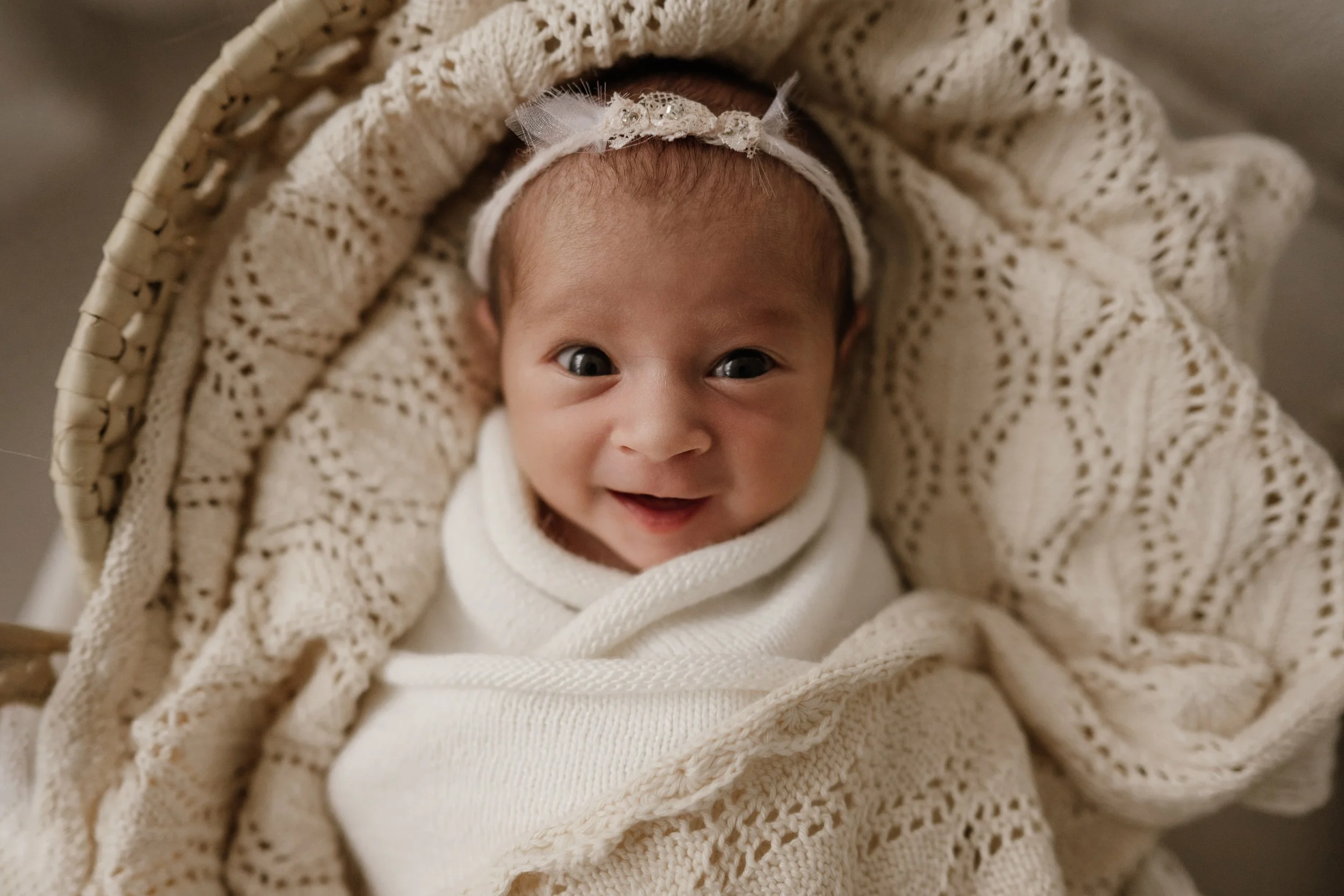 A smiling baby wrapped in a cream-colored knitted blanket, wearing a white headband with lace, lying in a wicker bassinet.