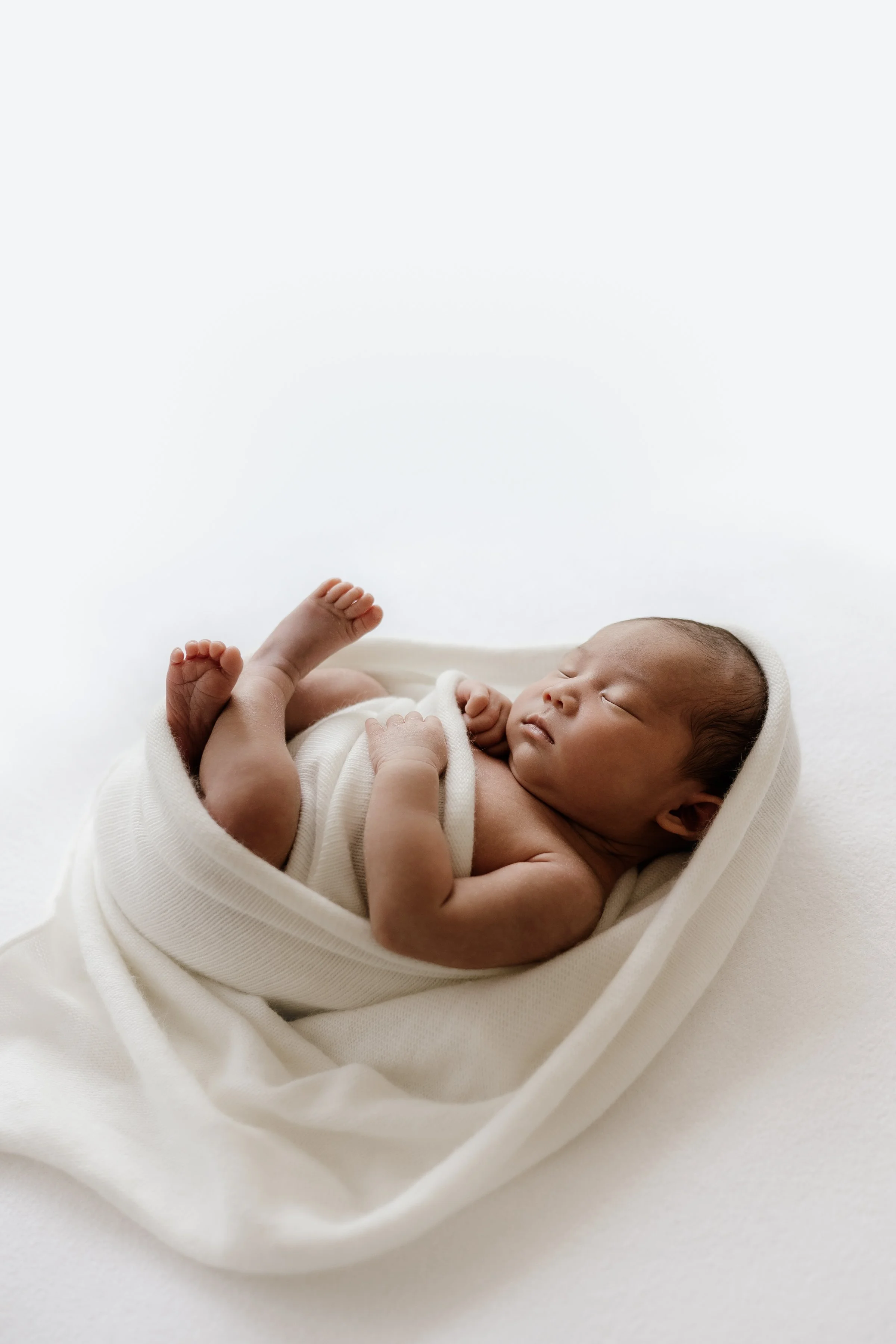 Newborn baby sleeping wrapped in white cloth on white background.