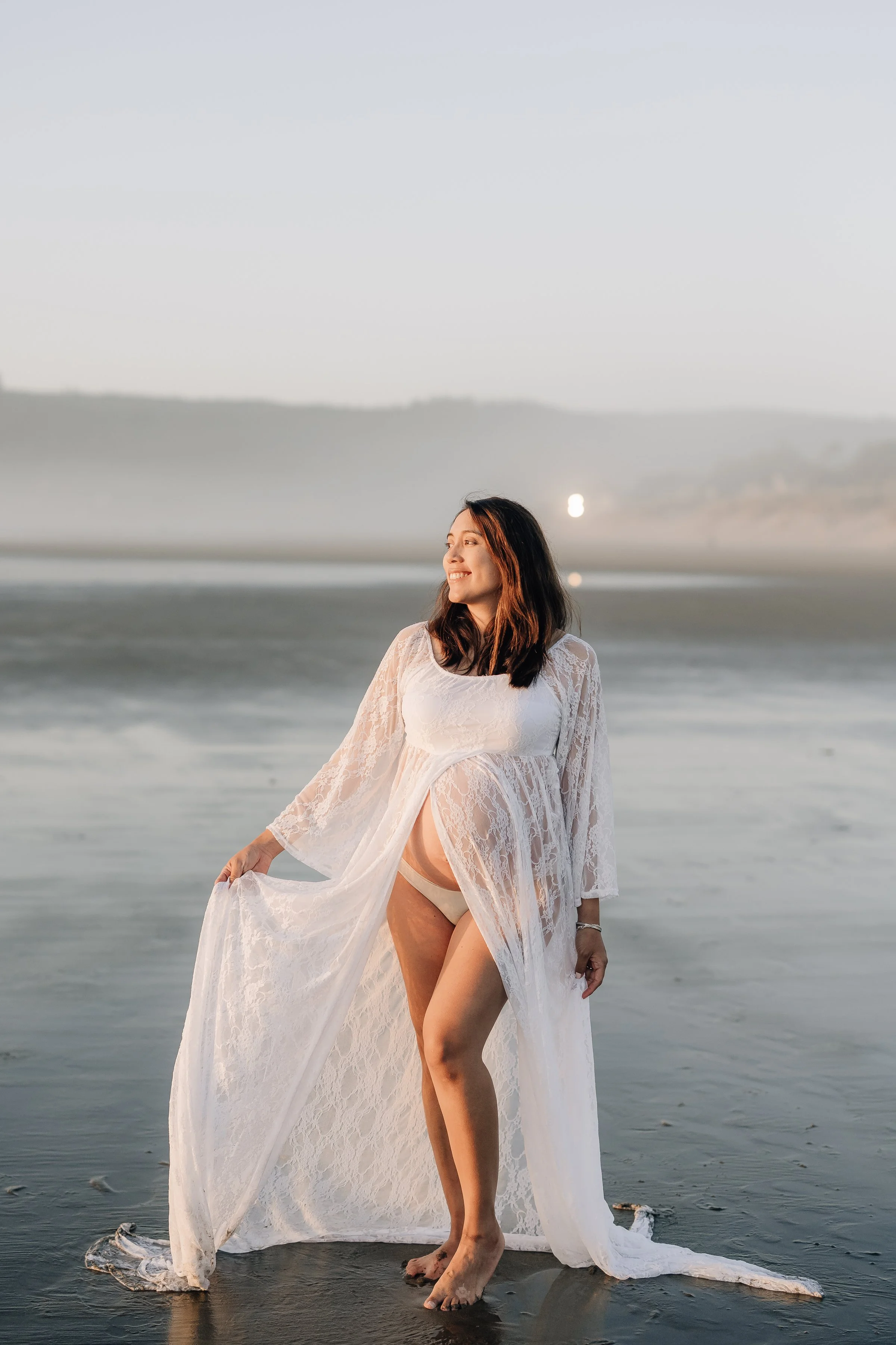 Woman in white lace dress standing barefoot in shallow water at the beach during sunset, smiling and looking to her left.