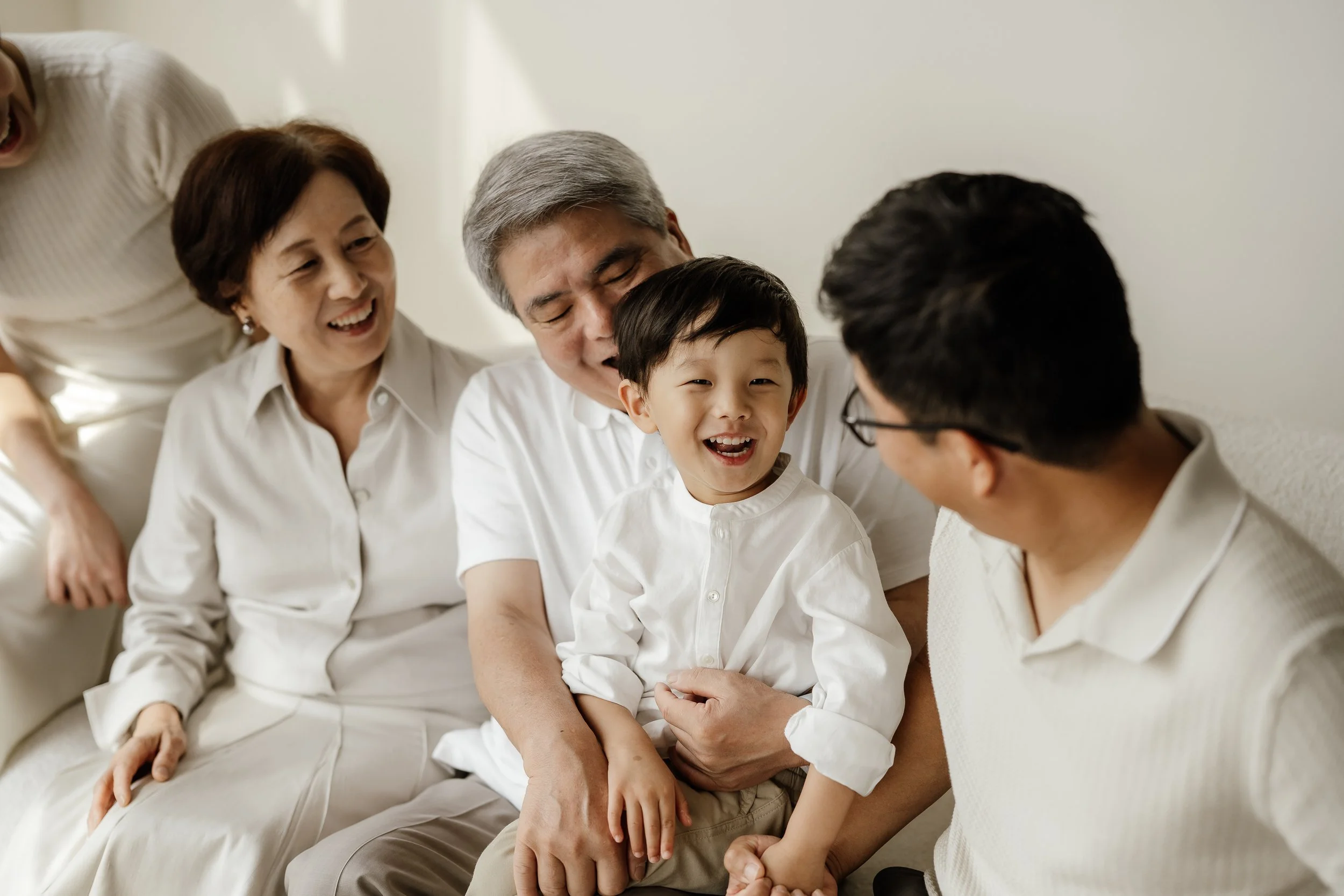 Smiling Asian family with grandparents and parents sitting together on a cream sofa in a room with bright lighting, engaging with a young boy.