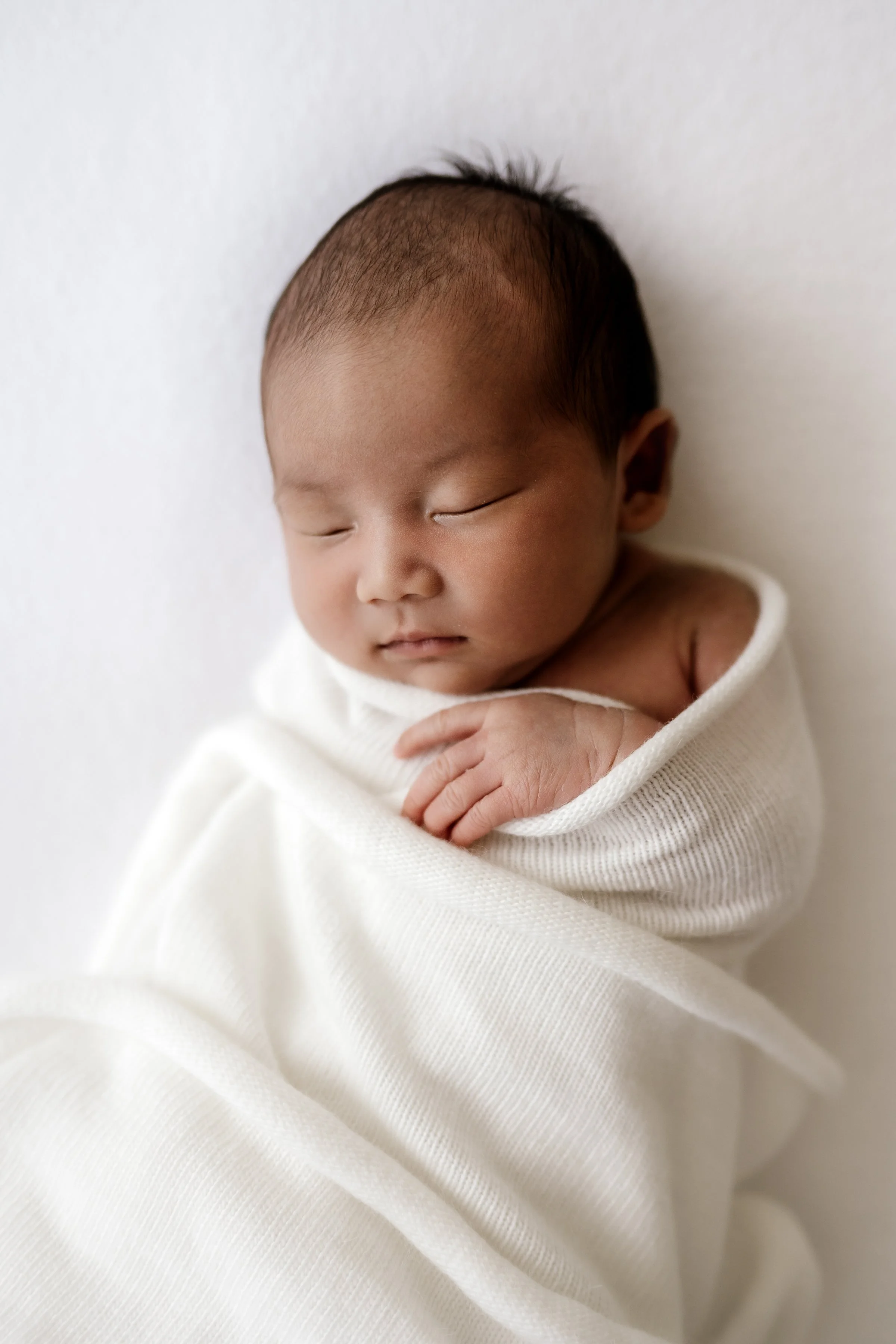 A sleeping newborn wrapped in a white blanket, lying on a soft white surface.