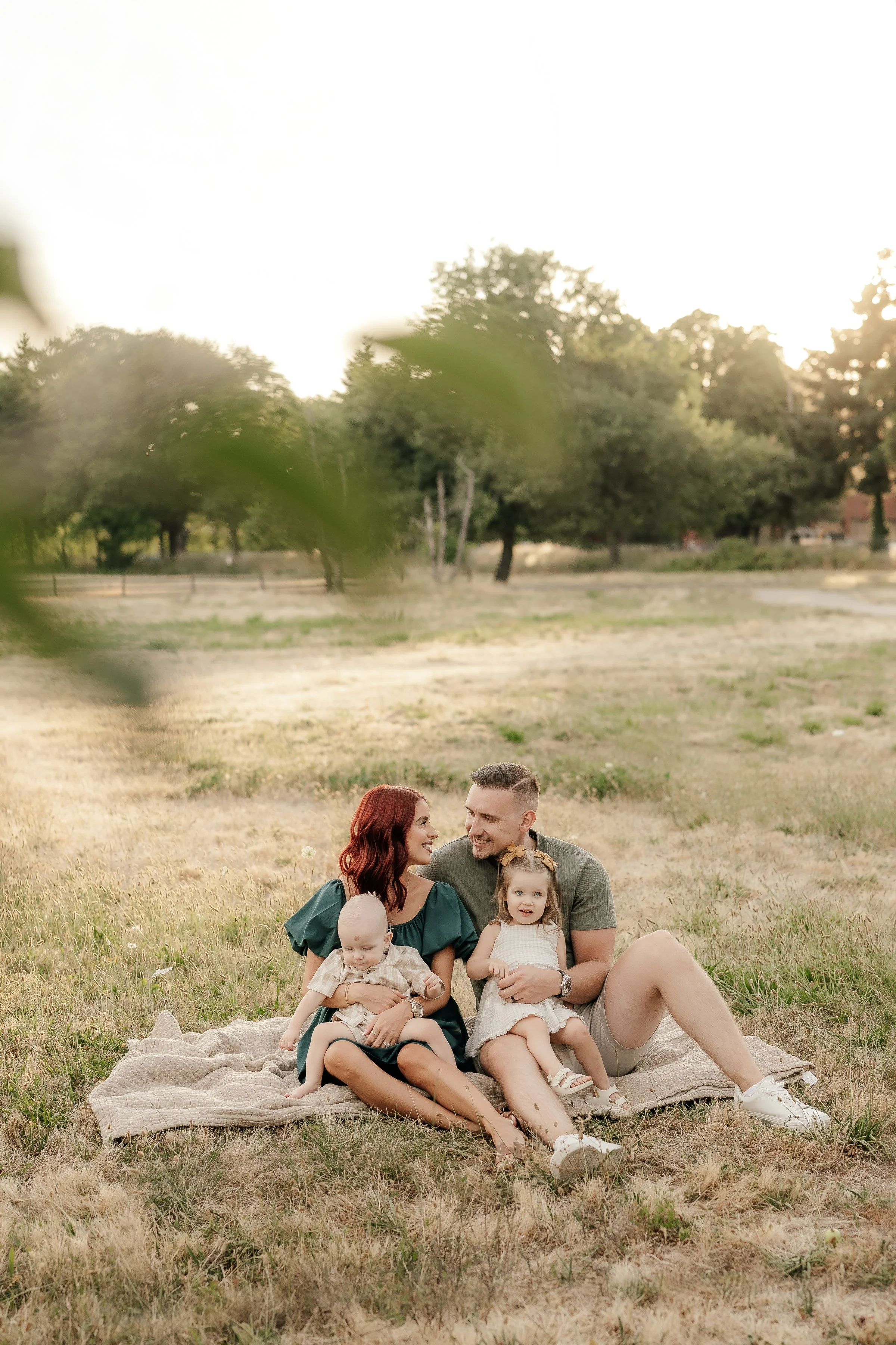 A family of four sitting on a blanket in a grassy park during sunset, with trees in the background.