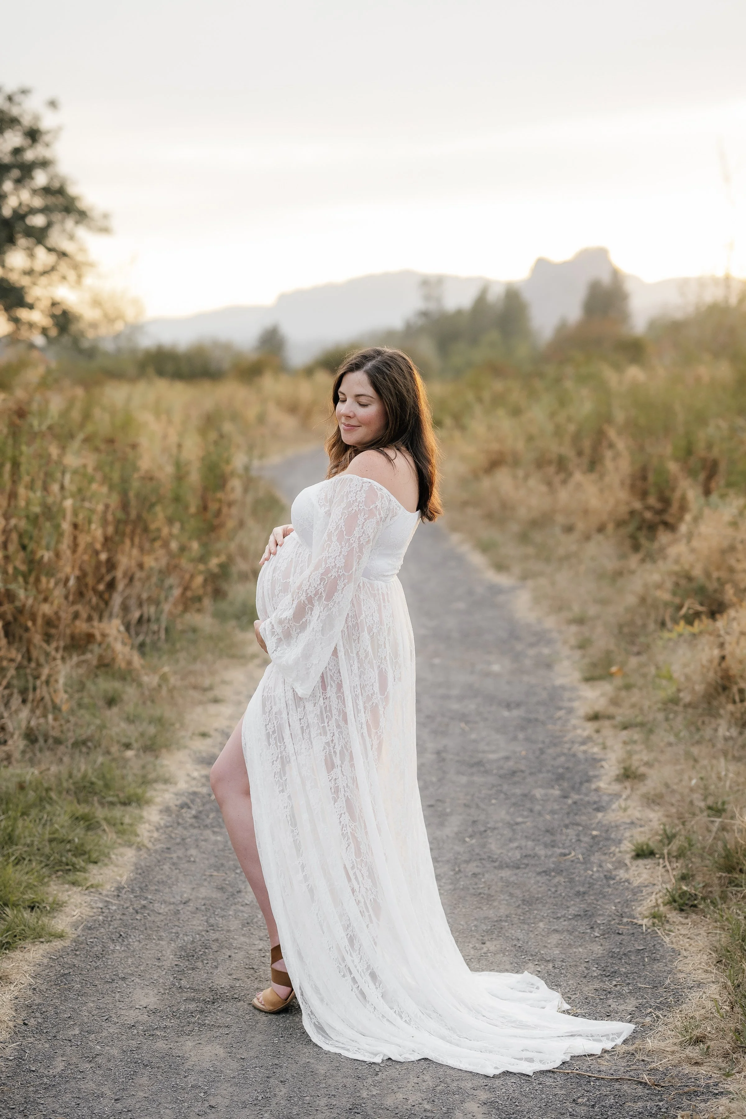 A pregnant woman in a long white lace dress standing on a dirt path in a natural, outdoor setting during sunset, holding her belly and smiling with her eyes closed.