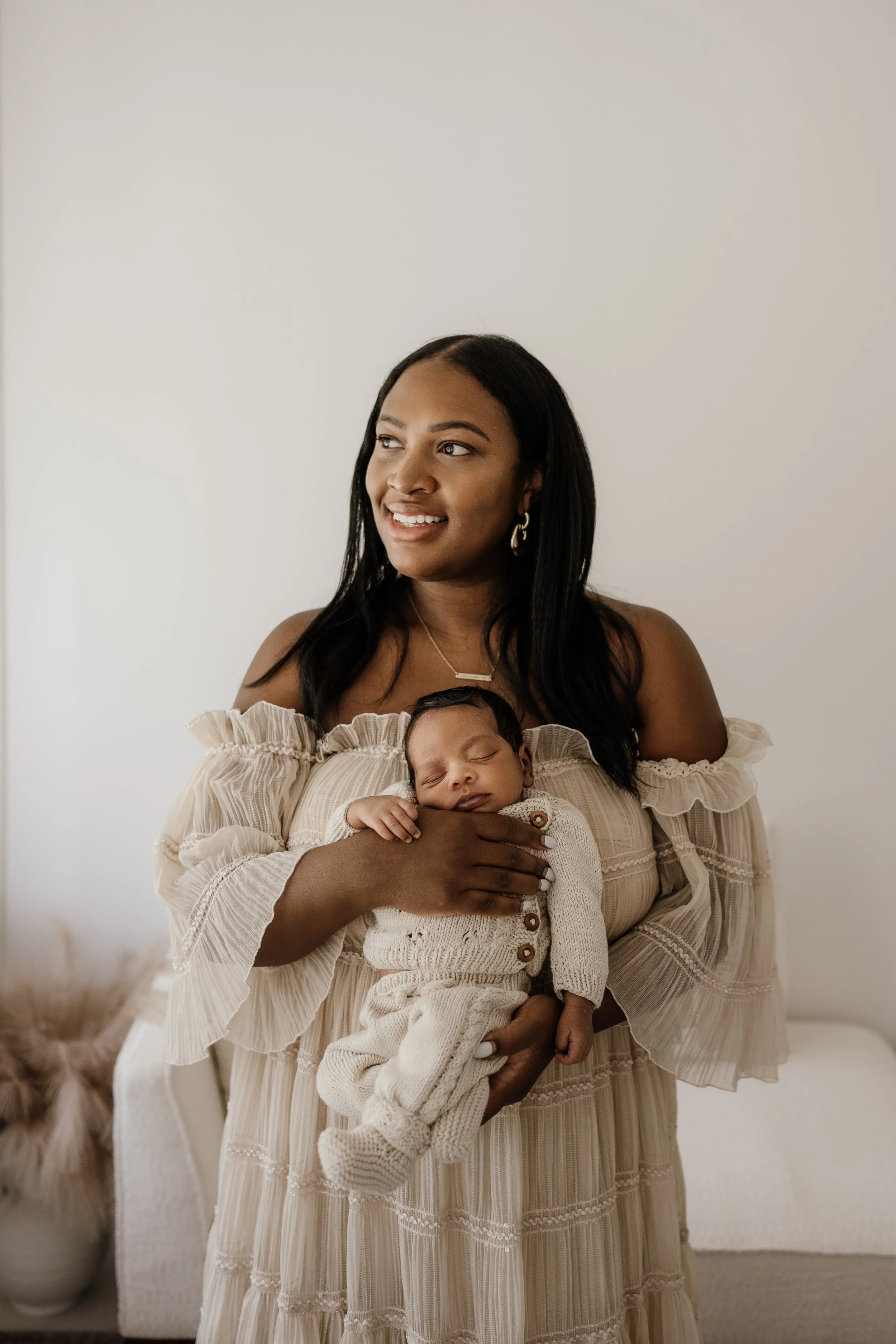 A woman holding a sleeping baby in a cream-colored outfit inside a room with a white wall and a floral vase in the background.