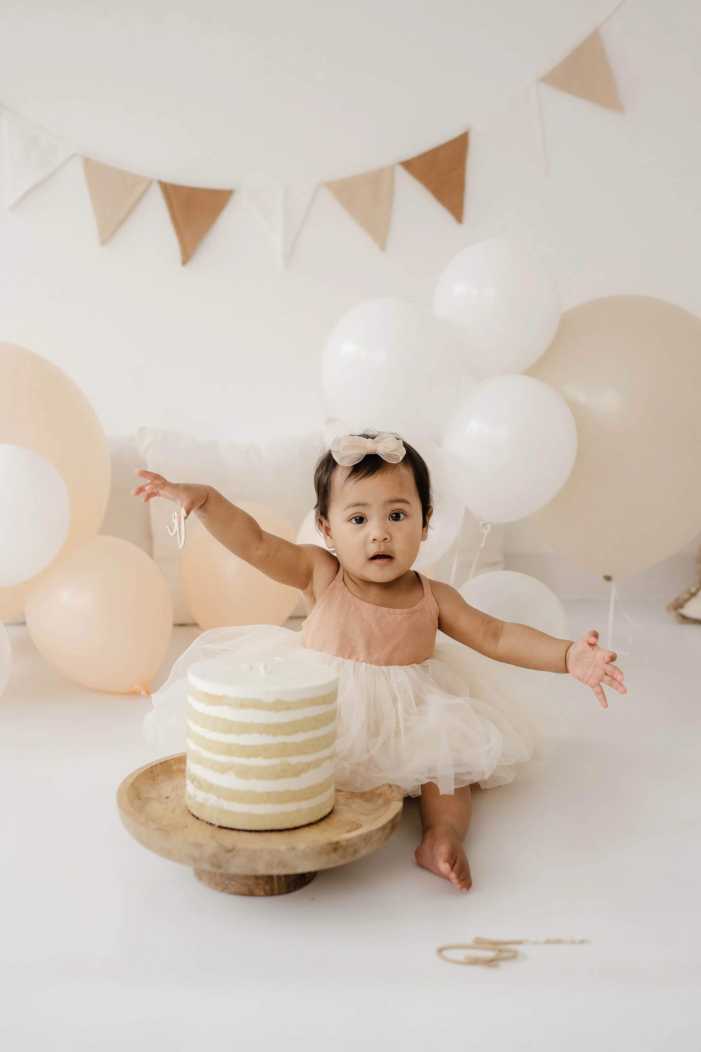 A young child in a peach-colored dress and tulle skirt sitting on the floor with arms outstretched, surrounded by beige and white balloons, with a small layered cake on a wooden cake stand in front of her, and beige and white pennant banners hanging