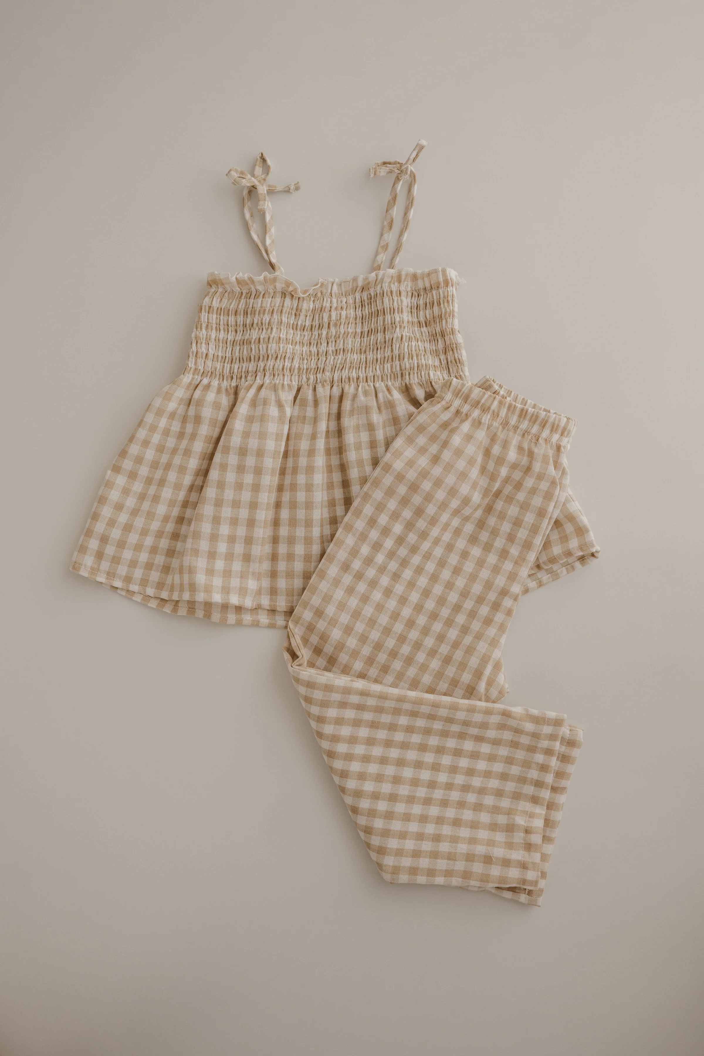 A set of beige and white checkered pajamas, including a sleeveless top with tied shoulder straps and matching pants, laid out on a white surface.