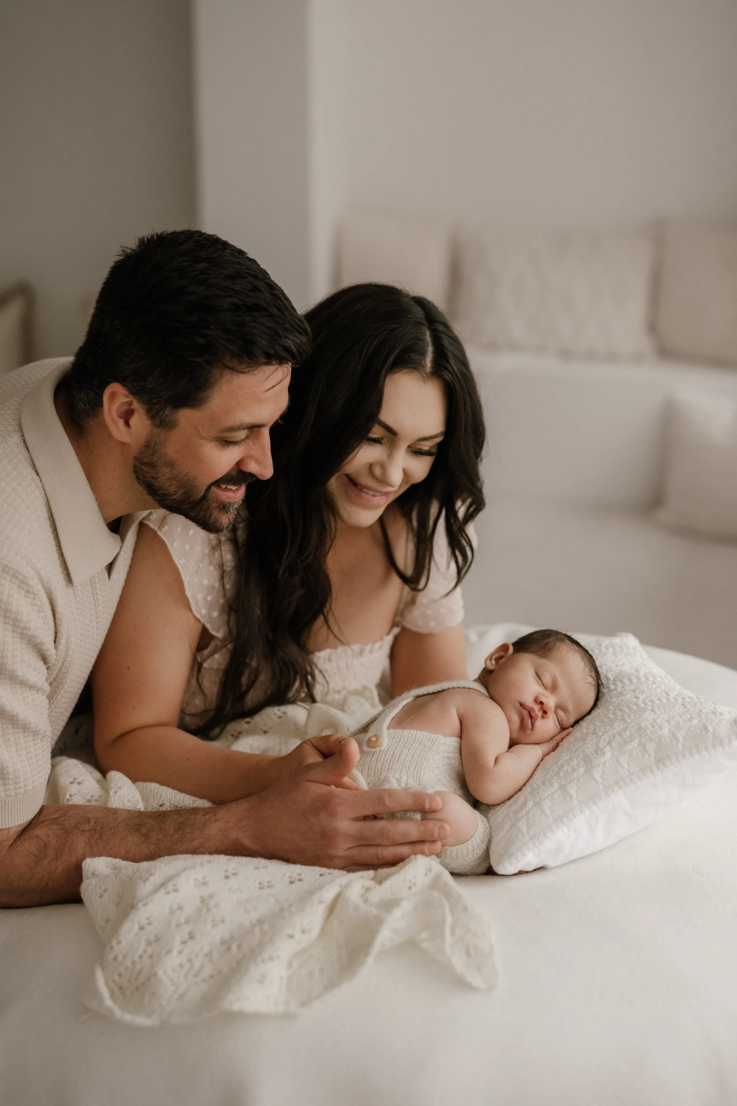 A young couple smiling and looking at their sleeping baby on a white bed.