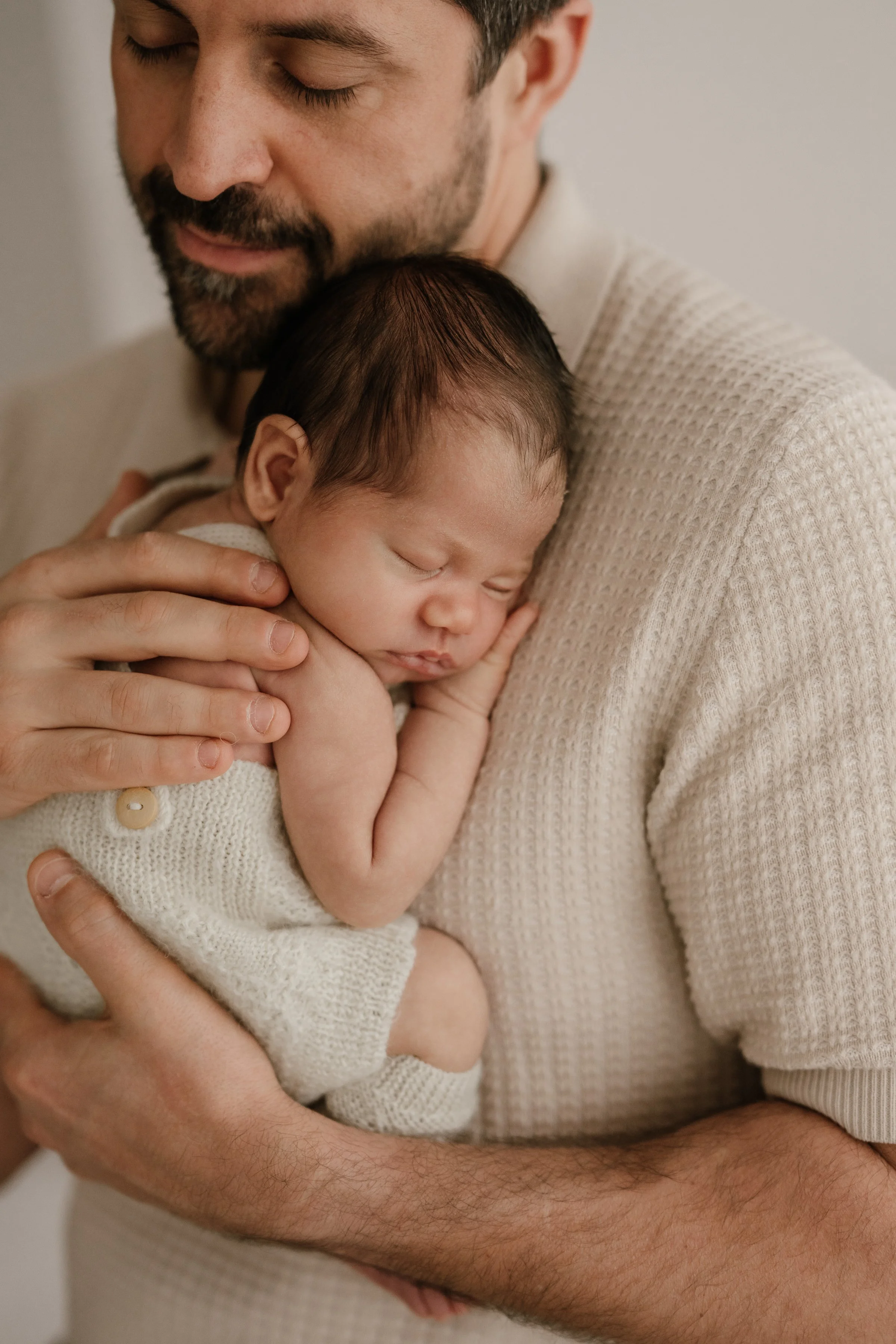A man with a beard gently holds a sleeping baby against his chest, both wearing neutral-colored clothing, with a calm and tender expression.