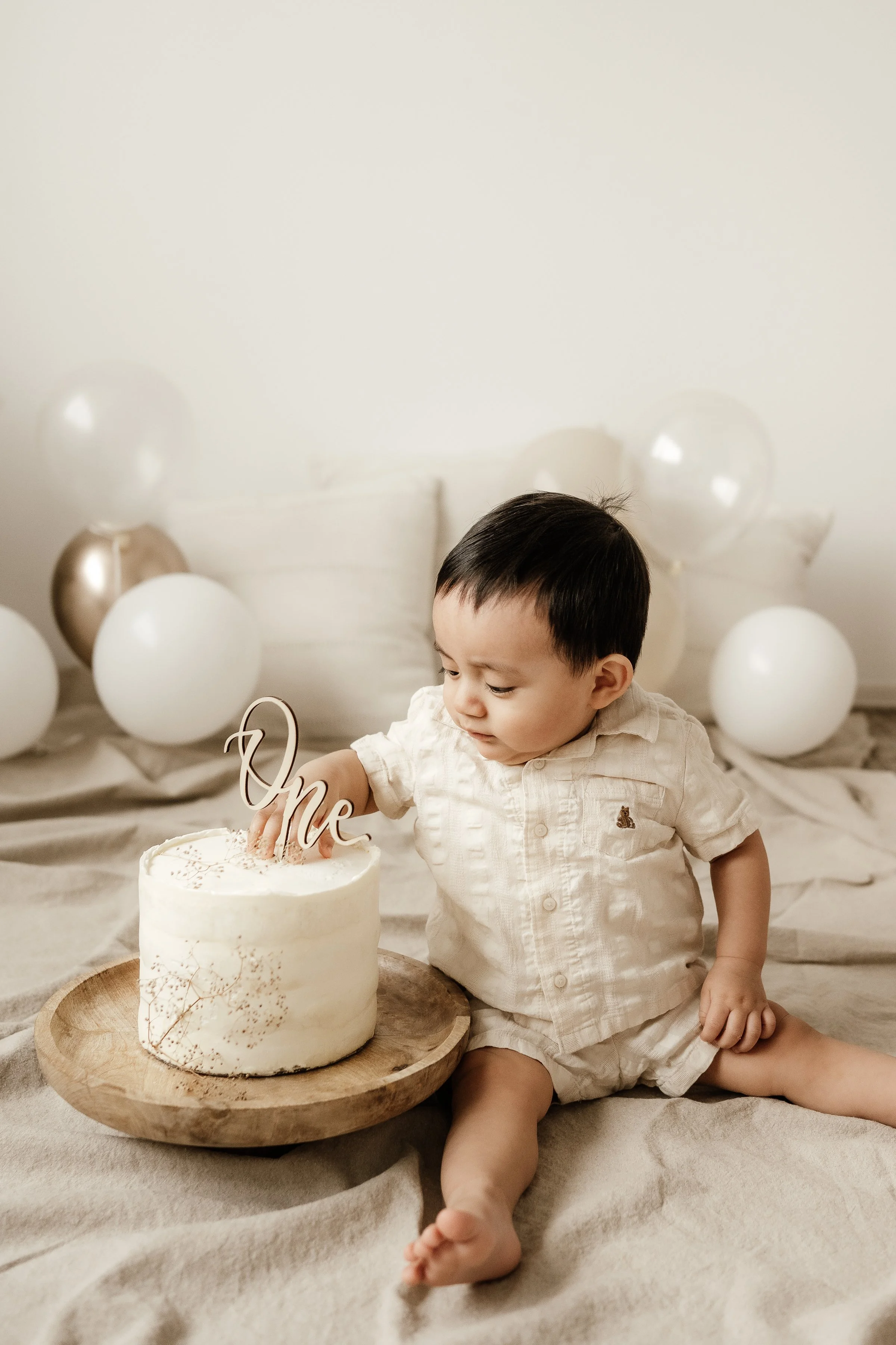Cute baby boy in a light-colored shirt and shorts sitting on a bed, reaching for a small white birthday cake with a decorative topper that says 'One.' Balloons are arranged in the background for a birthday celebration.