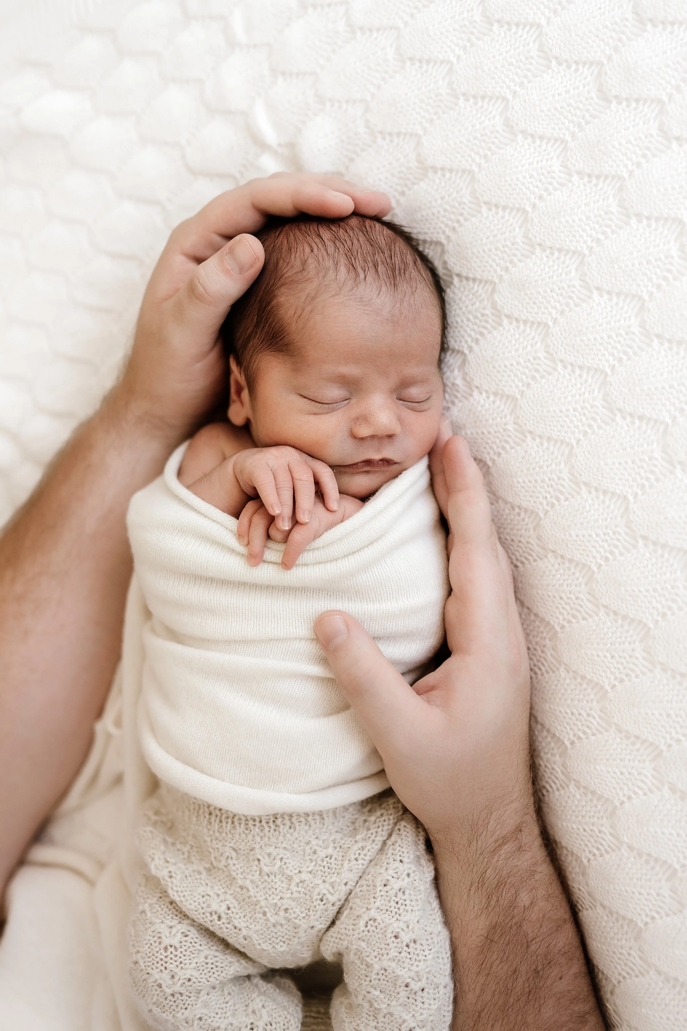 A newborn baby peacefully sleeping, held gently by adult hands, on a textured cream-colored blanket, wrapped in a white cloth.