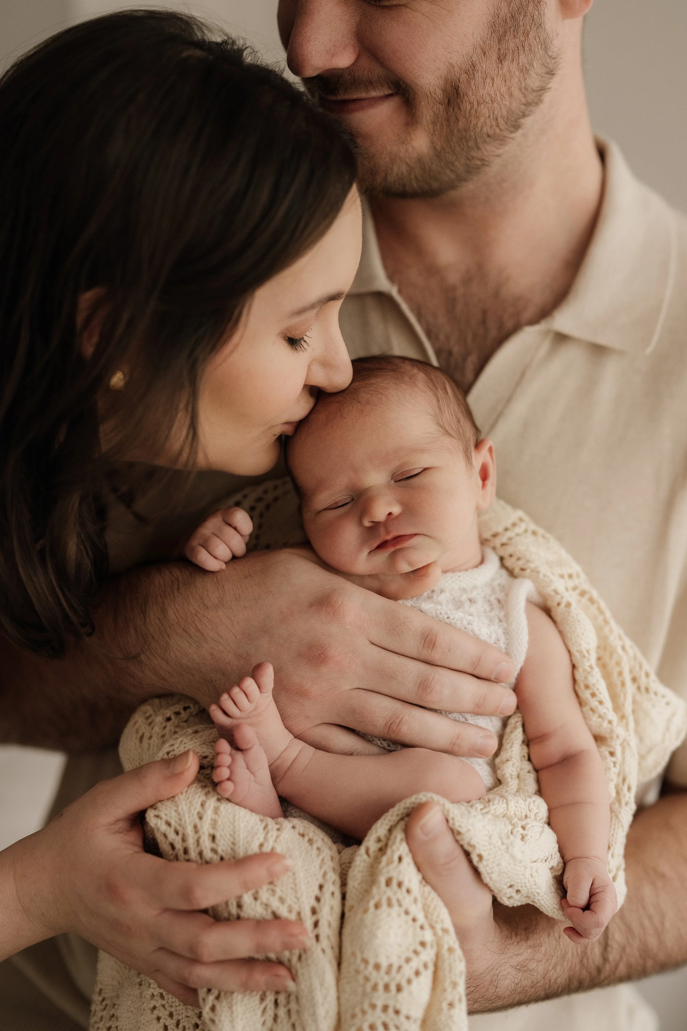 A woman and man holding a newborn baby, with the woman kissing the baby's forehead. The baby is wrapped in a knitted cream-colored blanket and has a peaceful expression.