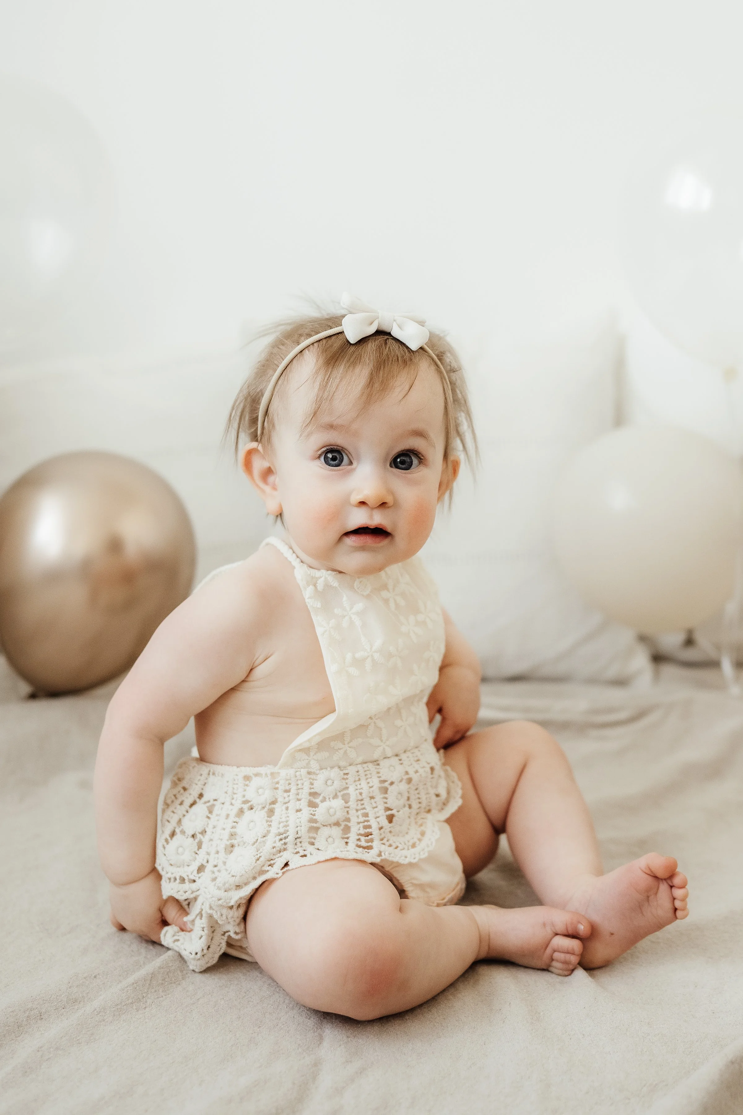 A baby girl with a bow headband sitting on a bed, surrounded by white and gold balloons, wearing a cream-colored dress with lace details.