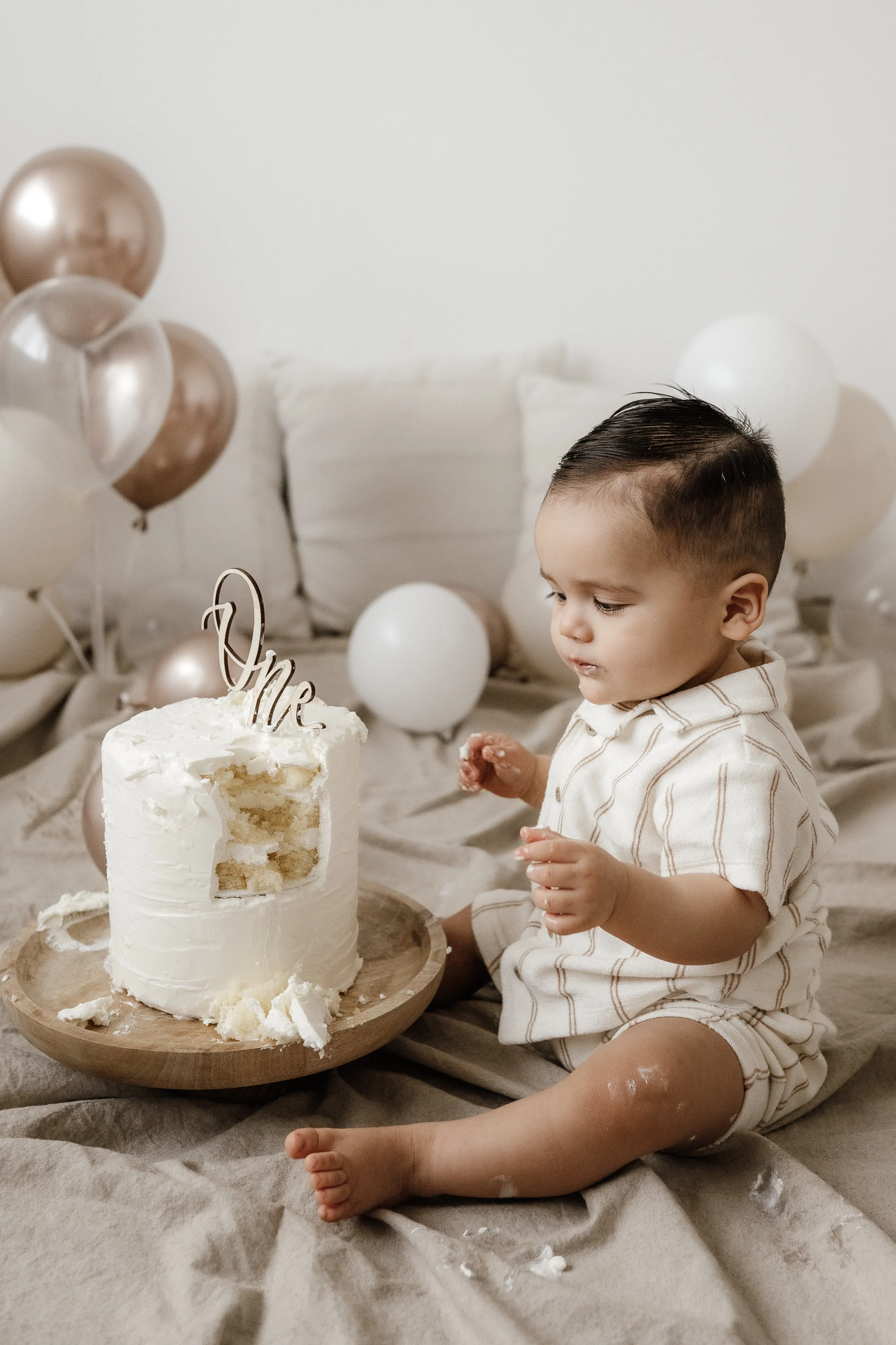 A young child celebrating their first birthday with a partially eaten white frosted cake on a wooden tray, surrounded by white and gold balloons, sitting on a beige blanket with beige pillows in the background.