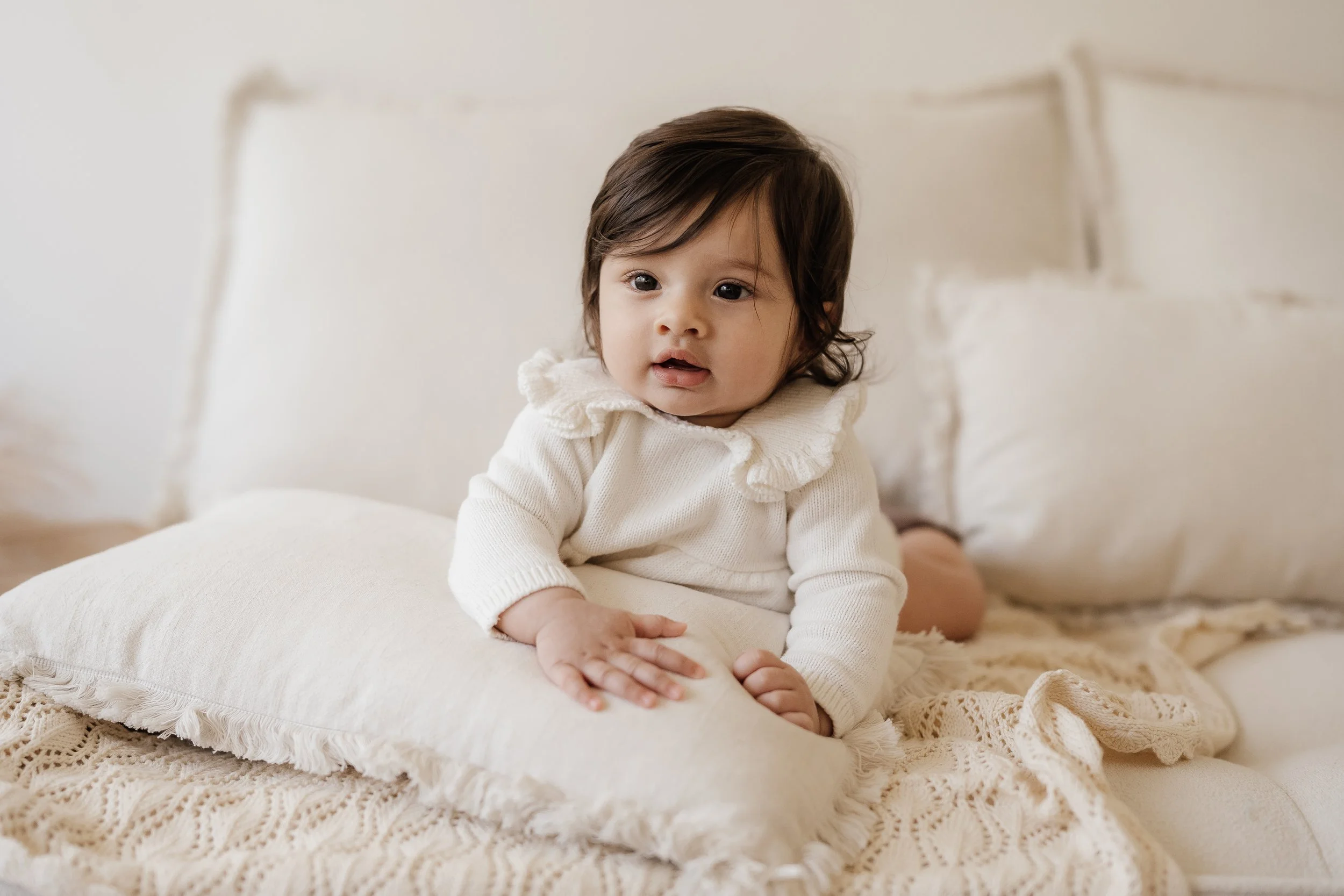 A young child with dark hair, sitting on a bed, wearing a white sweater with ruffled collar, surrounded by cream-colored pillows and blanket.
