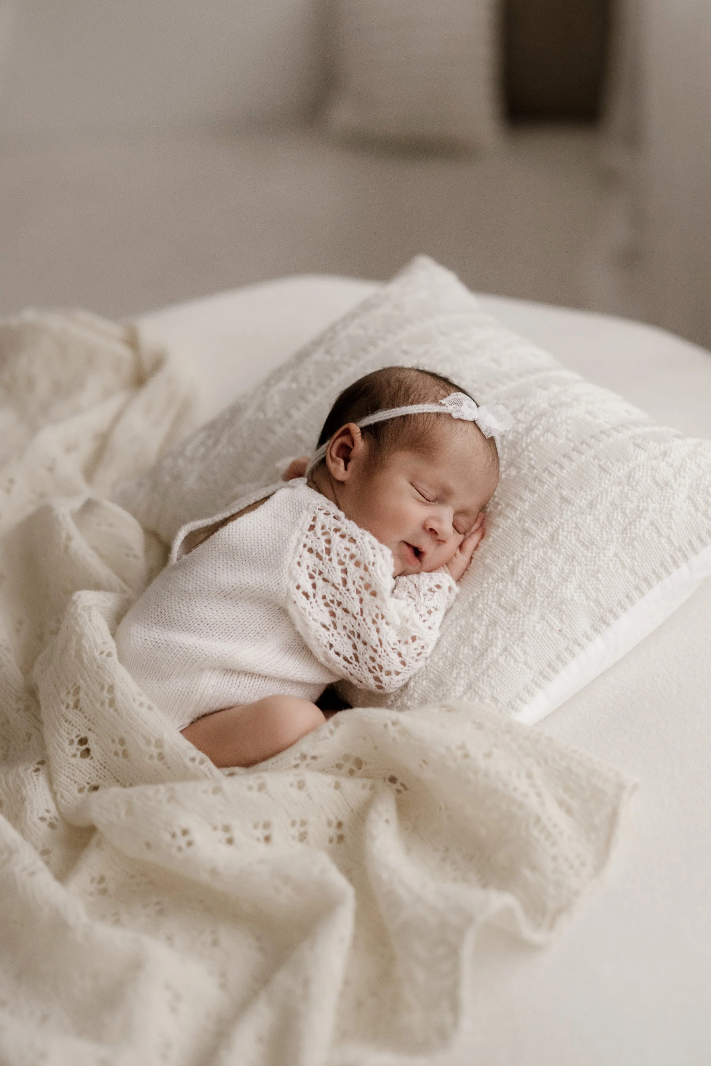 A newborn baby girl peacefully sleeping on a white bed, wrapped in a lacy cream-colored blanket, with a matching headband, lying on a soft white pillow.