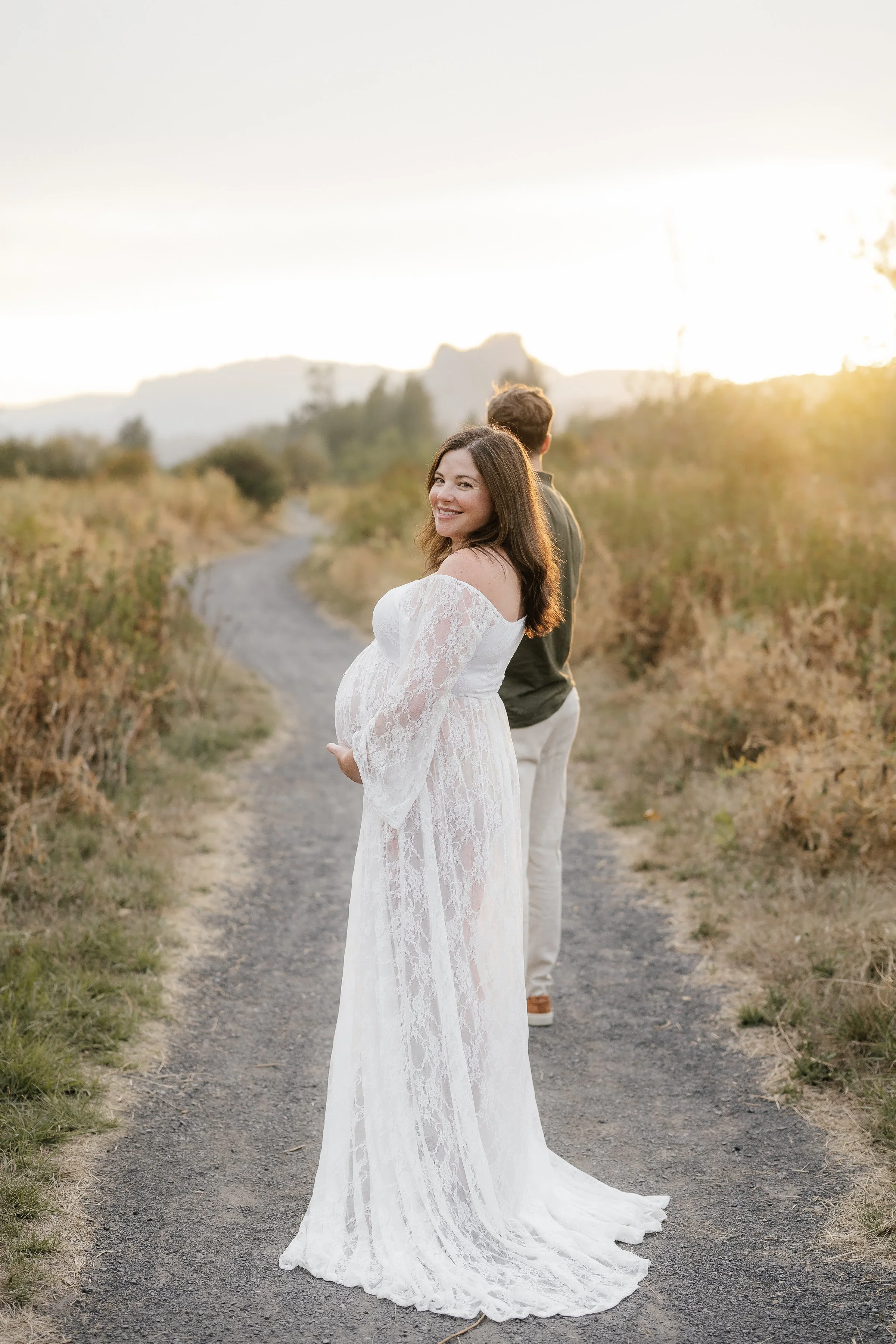 A smiling pregnant woman in a white lace dress standing on a dirt path in a rural area, with a man facing away in the background during sunset.