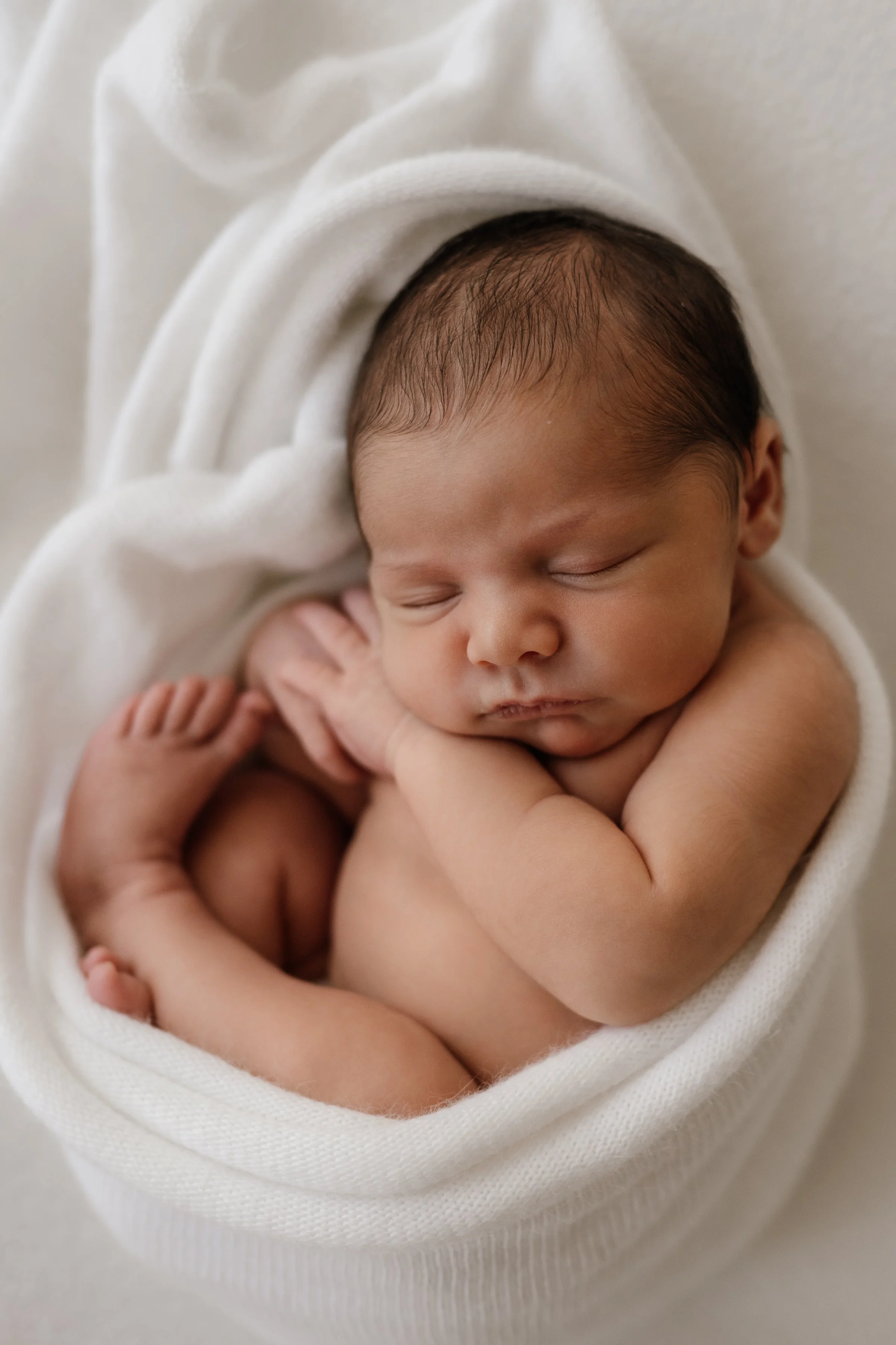 Sleeping newborn baby wrapped in a soft white blanket with arms folded, resting peacefully.